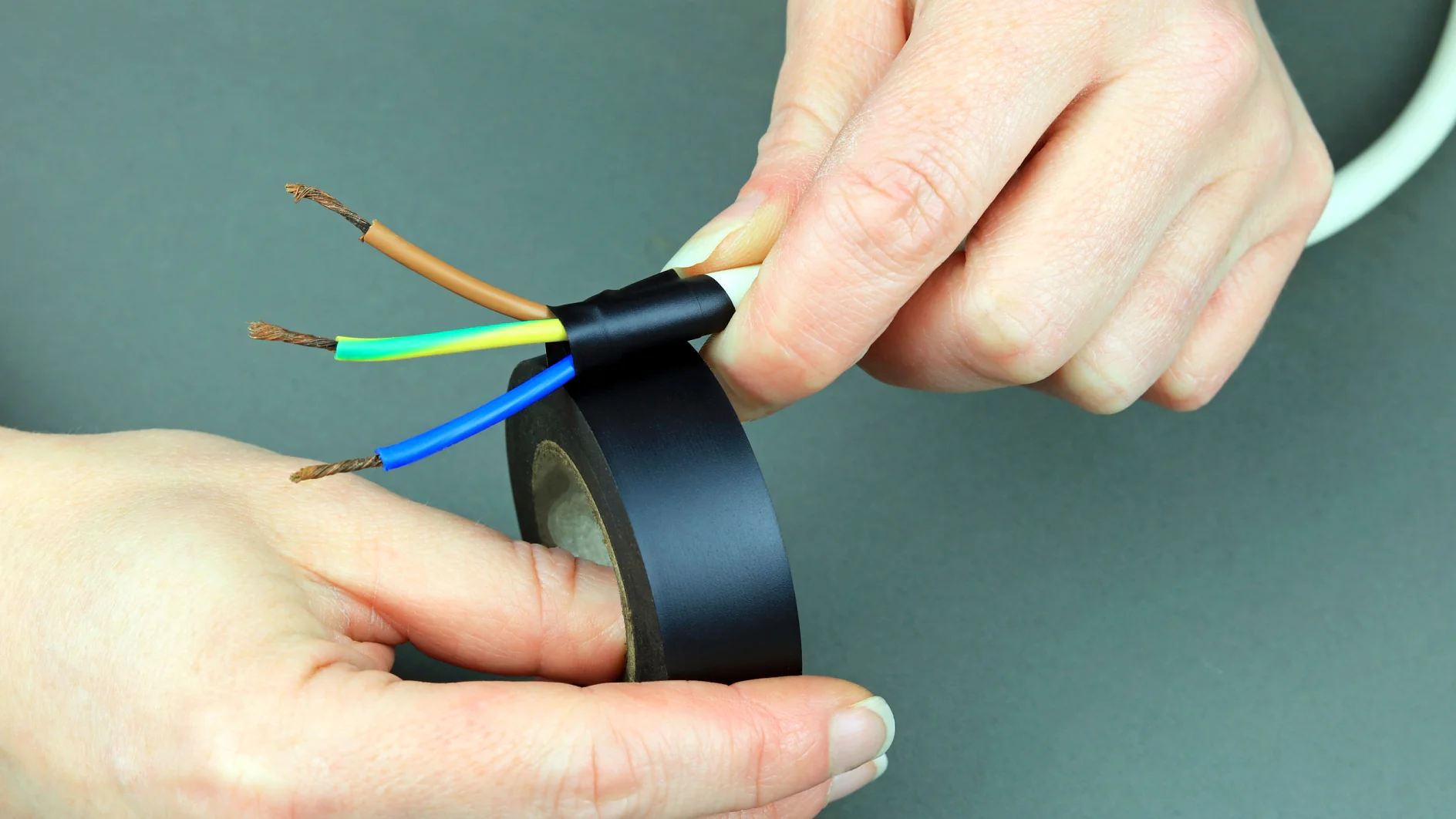 Close-up of hands wrapping black tesa tape around exposed wires with green, blue, and brown insulation. The background is a plain gray surface. (This text has been generated by AI)