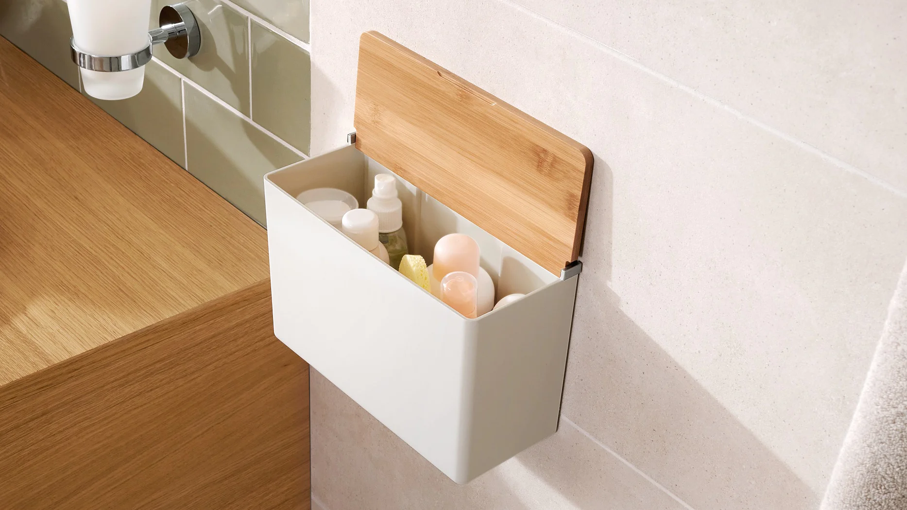 A white rectangular wall-mounted storage box with a wooden lid is open, revealing various bottles inside. It is attached to a bathroom wall using tesa tape, near a wooden countertop with a bowl sink and a tray holding jewelry. A towel hangs to the right. (This text has been generated by AI)