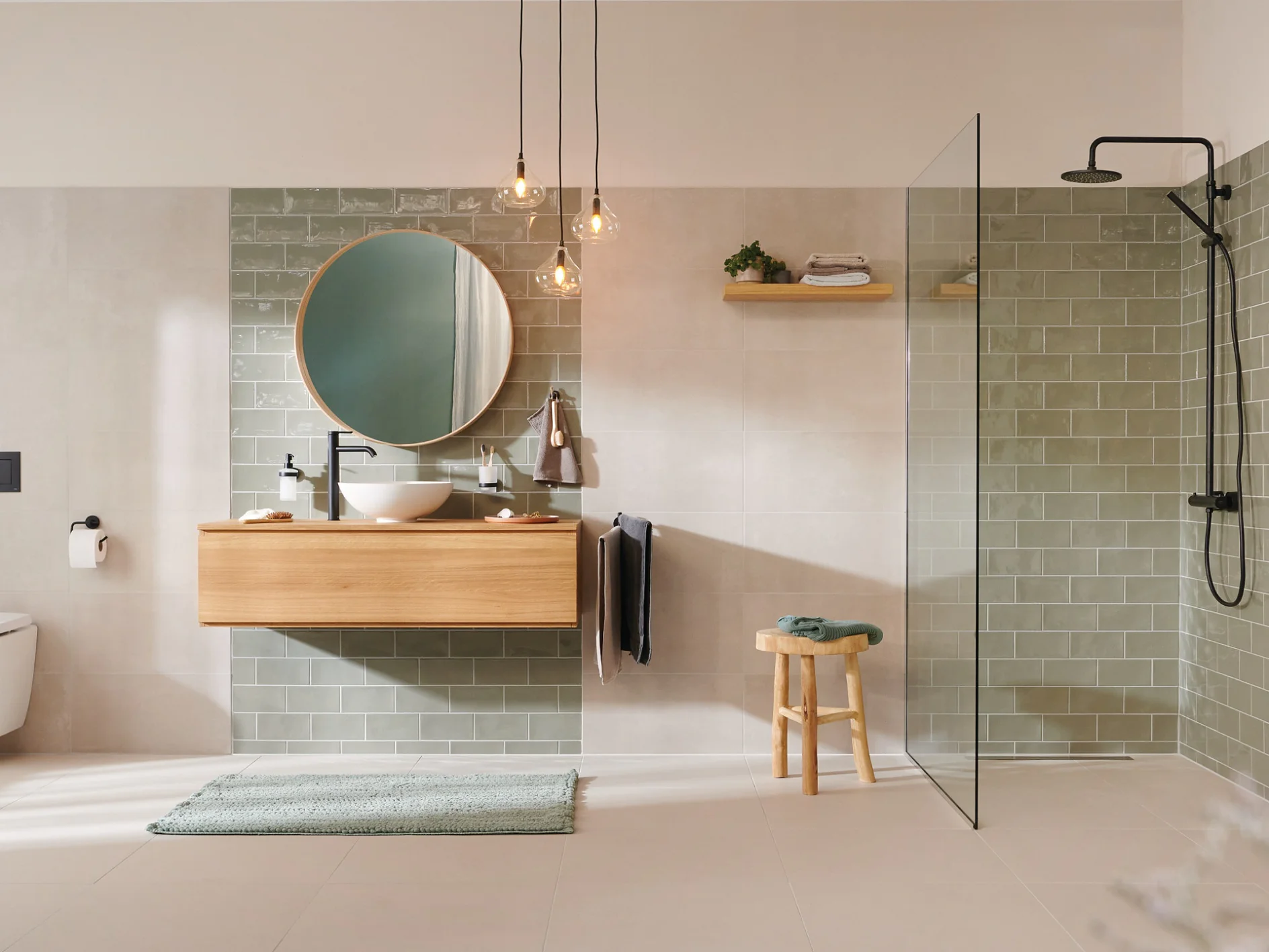A modern bathroom with beige tiles and a green tiled accent wall. Features include a round mirror above a wooden vanity with a white sink, a walk-in shower with black fixtures, a wall-mounted toilet, pendant lights, and a towel on a hook secured with tesa tape. (This text has been generated by AI)