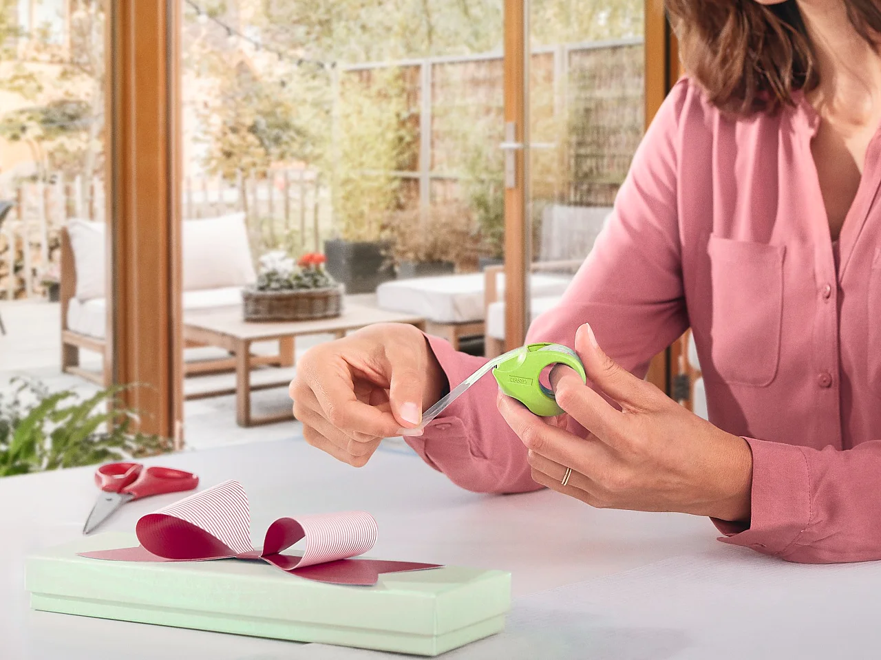 A woman in a pink blouse wraps a gift at a table. She is using green ribbon and pink wrapping paper, securing everything with tesa tape. The room has large windows with a view of trees and a patio. There is a gray couch and pillows in the background. (This text has been generated by AI)