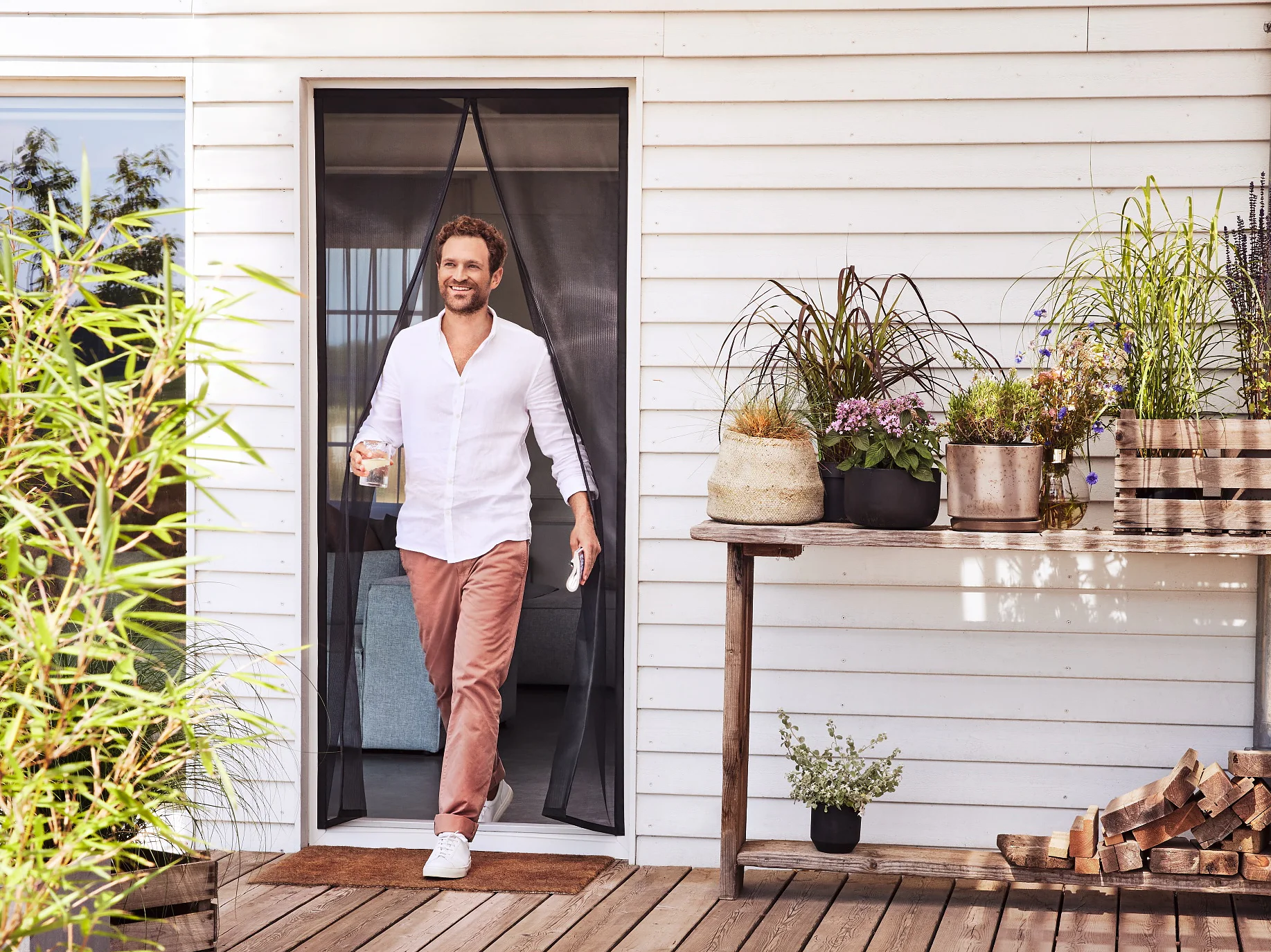 Un homme vêtu d'une chemise blanche et d'un pantalon rose franchit une porte pour accéder à une terrasse en bois, un verre à la main. La terrasse est agrémentée de plantes en pot, d'une pile de bois de chauffage et d'une table avec d'autres plantes. Le bâtiment a un revêtement blanc. (This text has been generated by AI)