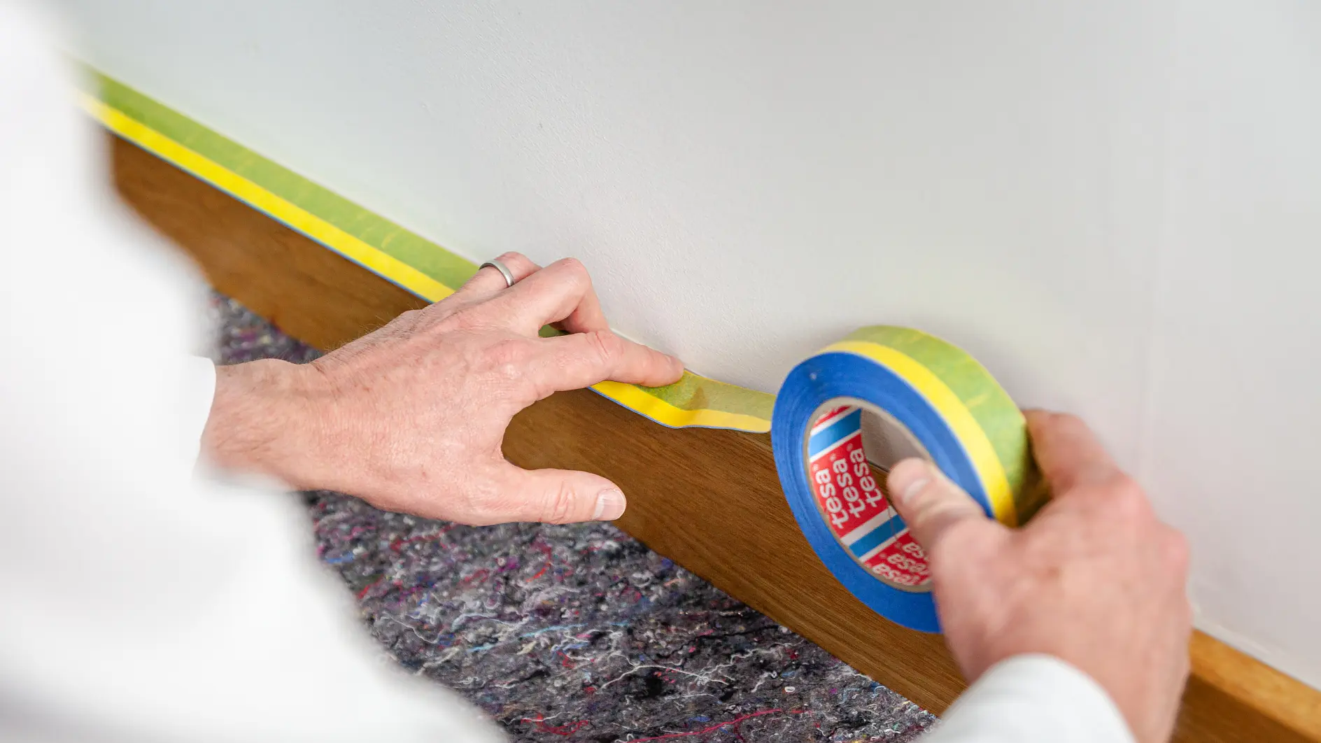Une personne applique du ruban tesa jaune le long du bord d’une plinthe en bois avant de peindre un mur blanc. Une bâche colorée protège le sol.