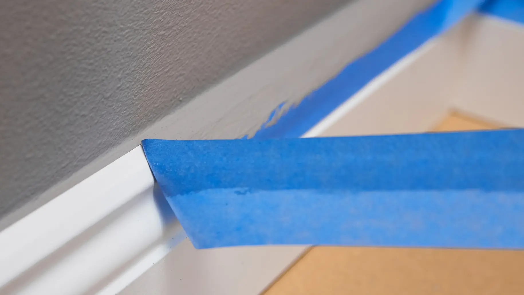 Une main retire un ruban tesa bleu de la plinthe blanche le long d’un mur gris, révélant une ligne de peinture nette et un sol protégé en dessous.
