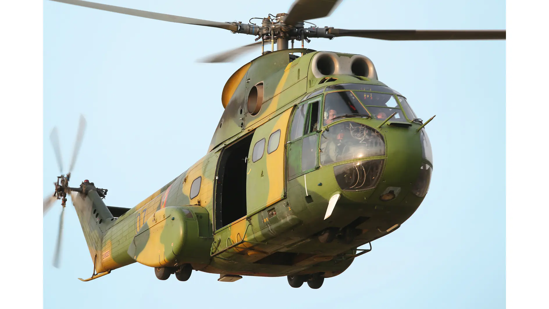 Un hélicoptère militaire vert camouflé, doté d’accents en ruban tesa, vole avec sa porte latérale ouverte, ses rotors tournant sous un ciel dégagé. (This text has been generated by AI)