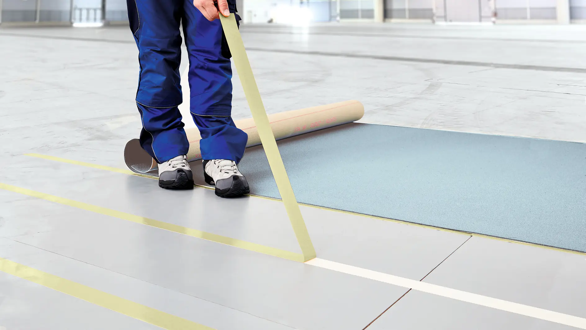 A person in blue pants and white sneakers is unrolling a large sheet of blue paper on a gray floor. They hold a roll of yellow tesa tape, applying it across the floor to create parallel lines. The background shows an empty, spacious room.