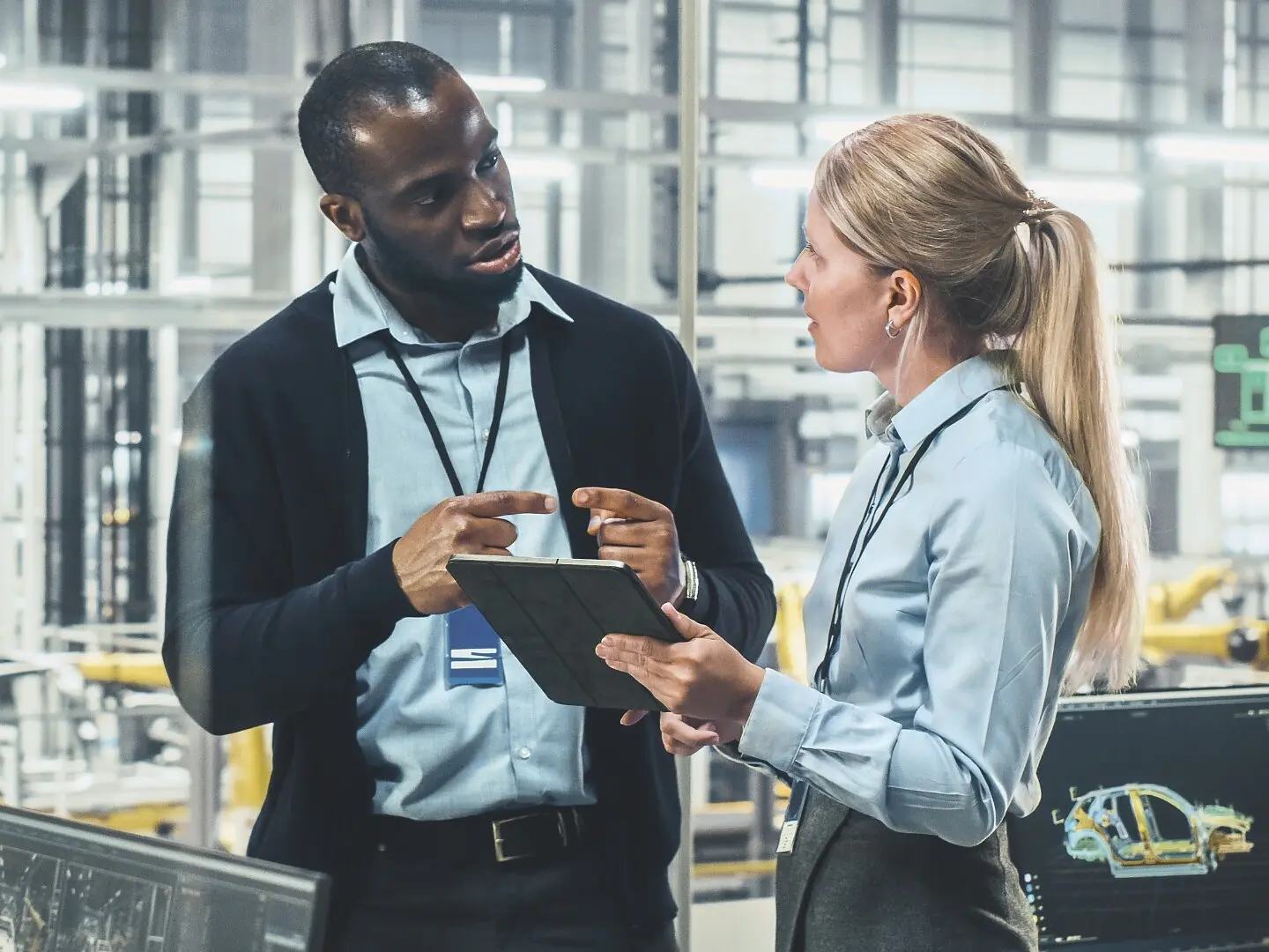 A man and a woman are standing in a modern industrial setting with tesa tape products in sight. The man is talking and gesturing while holding a notepad. They are both wearing business attire and ID badges. Various technical equipment and monitors are visible in the background, emphasizing the prominence of tesa tape in the environment. (This text has been generated by AI)