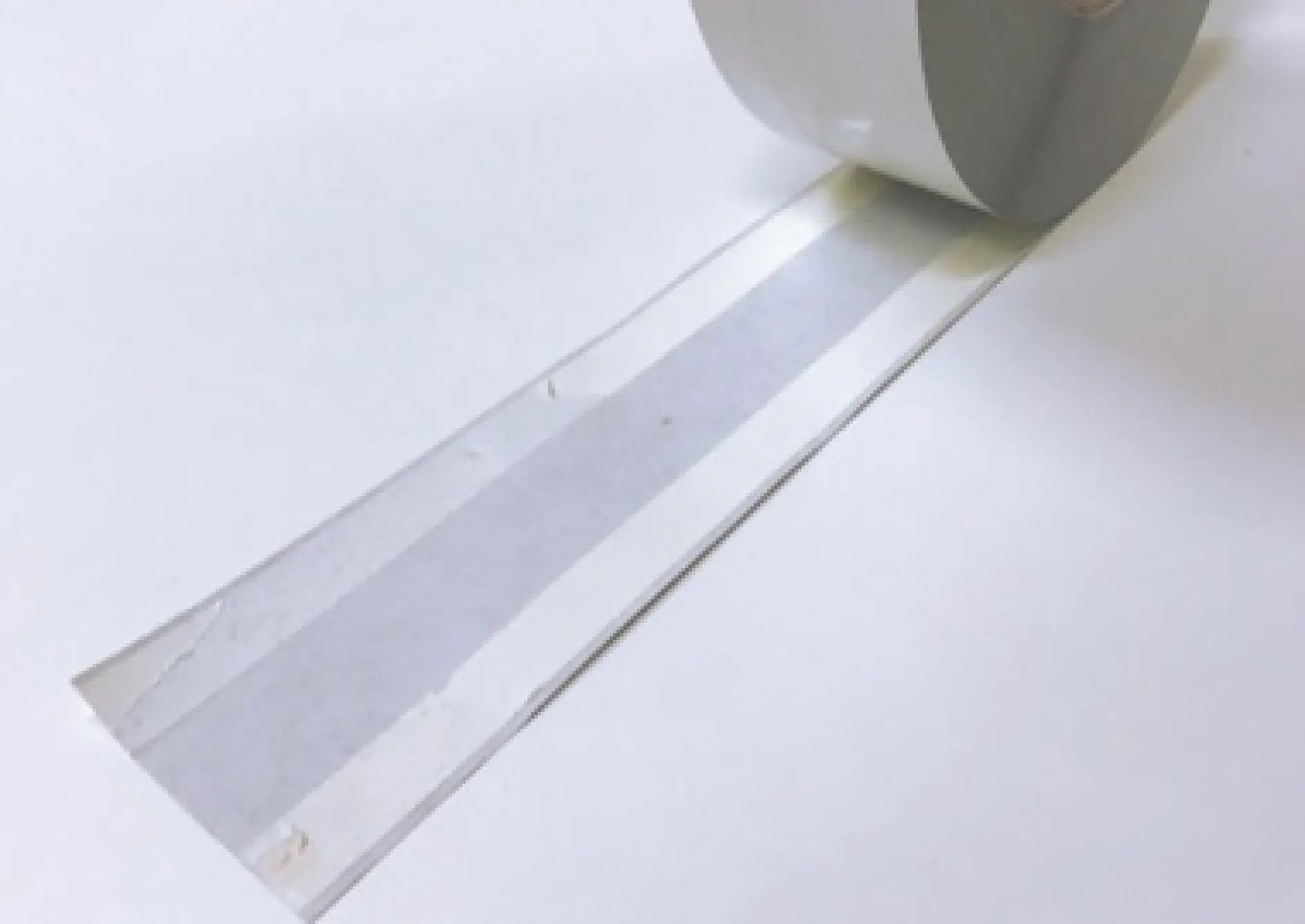 A roll of gray tesa tape is partially unfurled on a white surface. The tesa tape is smooth and reflective, showing a narrow, sticky strip on each side. (This text has been generated by AI)