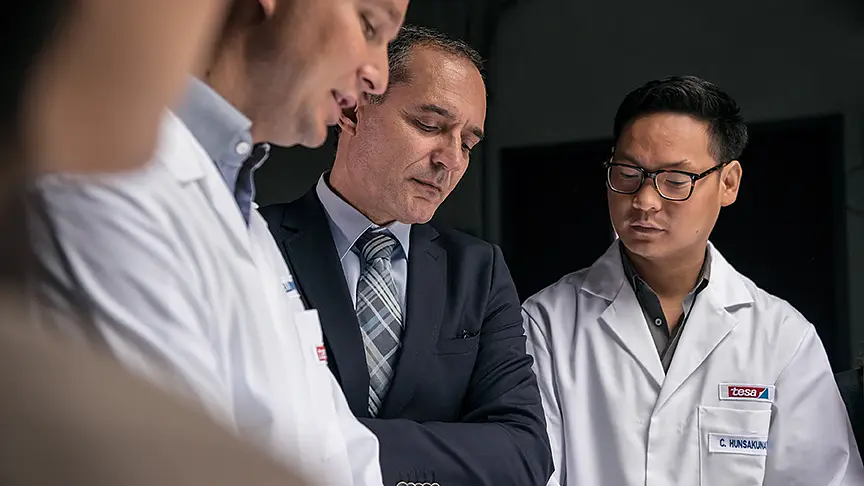Four men are gathered, three wearing white lab coats and one in a suit. They appear to be discussing something of interest or examining an object outside the frame, possibly involving tesa tape. The setting looks like a professional or laboratory environment. (This text has been generated by AI)