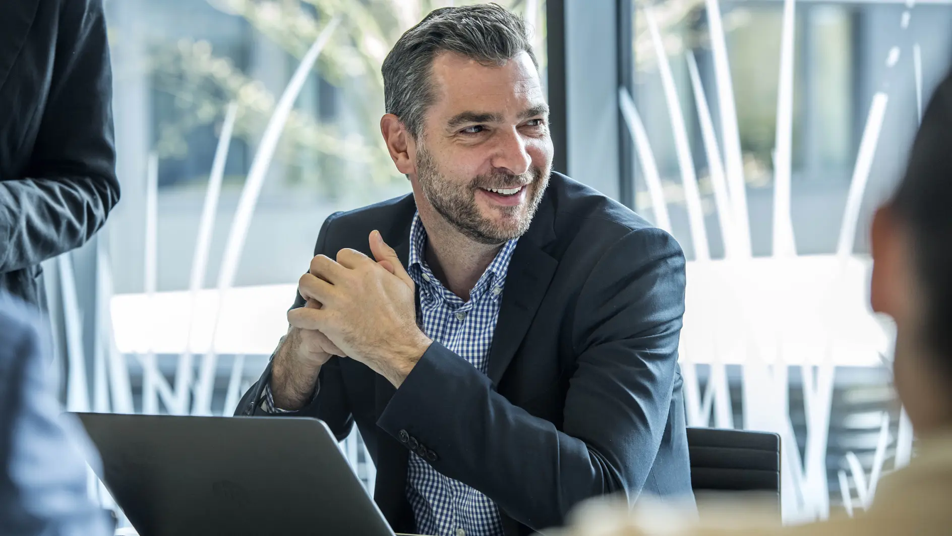 A man with graying hair and a beard is sitting at a desk, smiling, with his hands clasped. He is wearing a dark suit jacket over a checkered shirt. A laptop is open in front of him. Other blurred figures are visible in the background. (This text has been generated by AI)