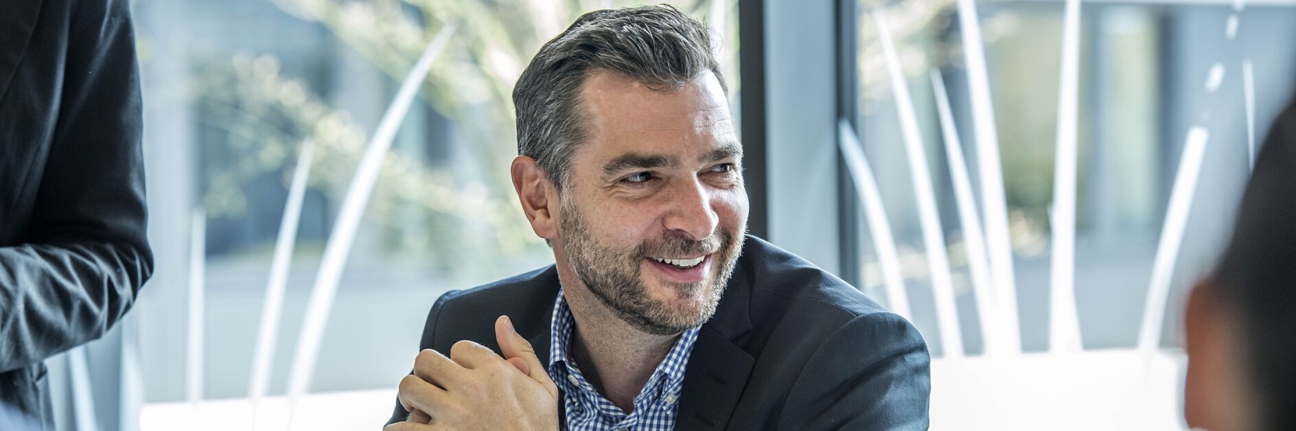 A man with graying hair and a beard is sitting at a desk, smiling, with his hands clasped. He is wearing a dark suit jacket over a checkered shirt. A laptop is open in front of him. Other blurred figures are visible in the background.