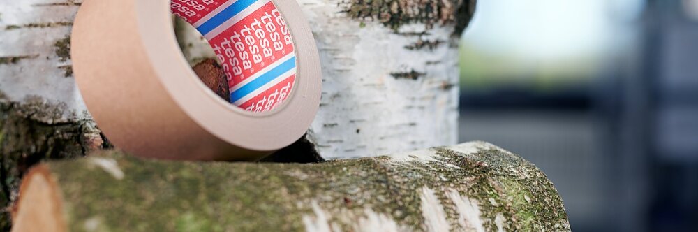 A roll of brown adhesive tesa tape is placed on a moss-covered log with other birch logs in the background. The scene is lit in natural light, emphasizing the texture of the tesa tape and the logs.