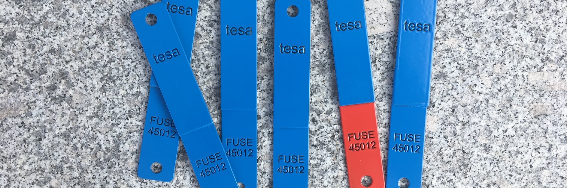 Seven blue metal strips with hole-punched ends, labeled with tesa and FUSE 45012, are laid flat on a speckled gray surface. Two strips have red ends.