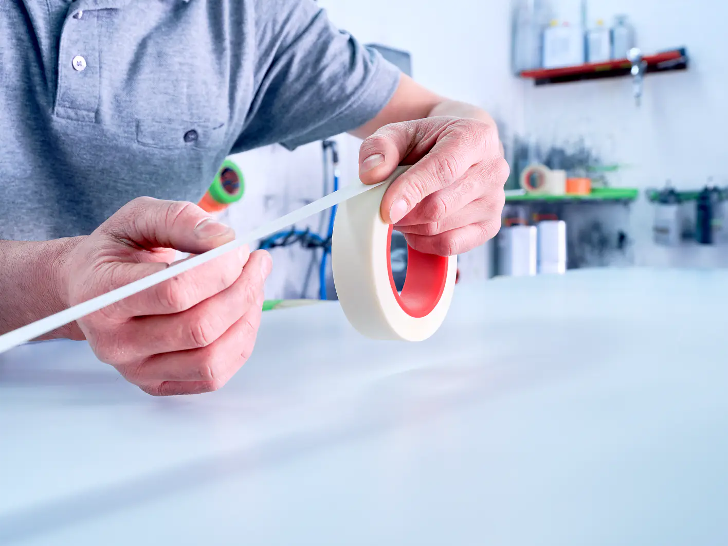 A person in a gray shirt holds a roll of tesa tape in one hand and pulls out a strip with the other hand. The background includes shelves with various containers and tools. (This text has been generated by AI)