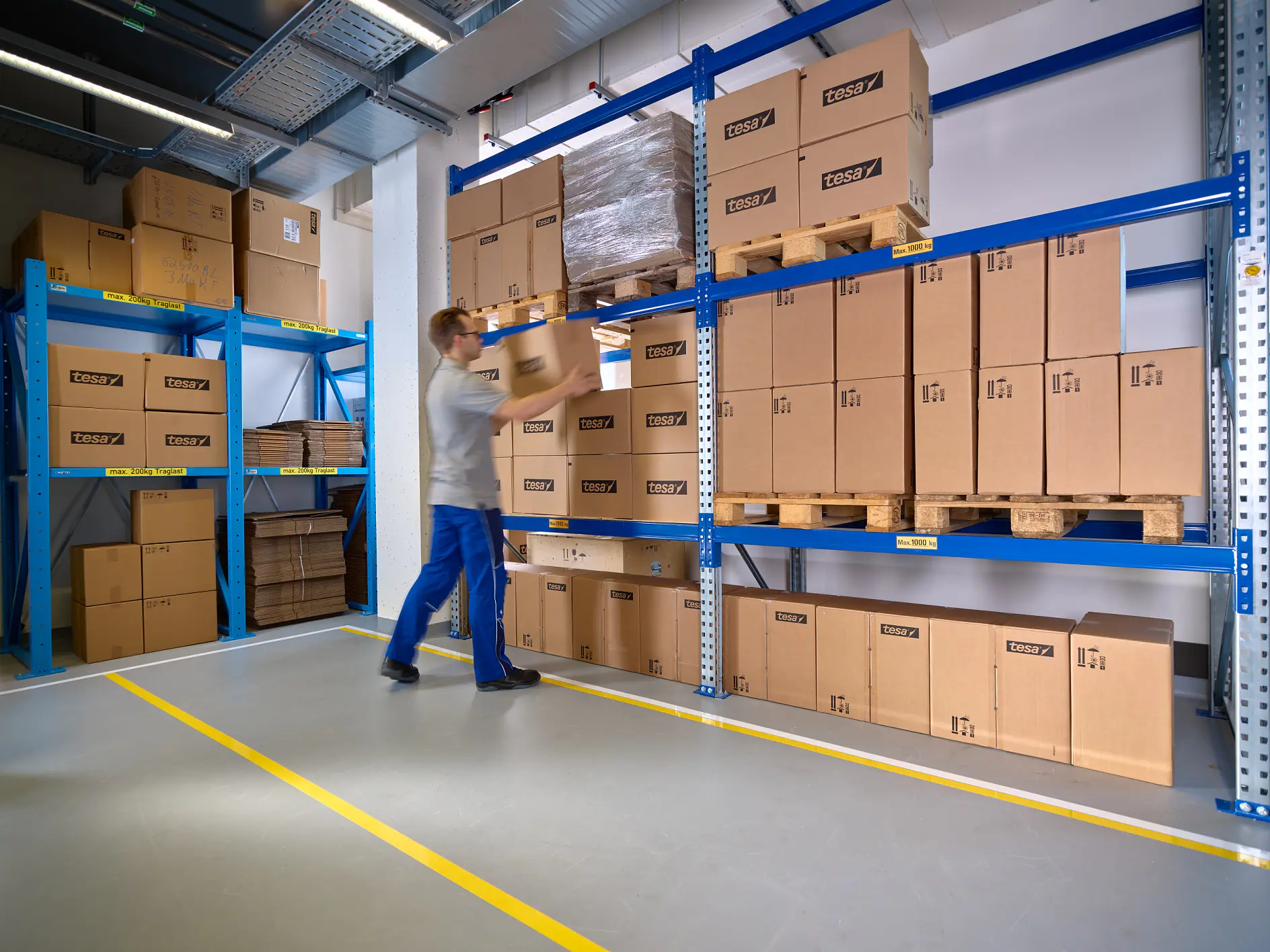 A worker in a gray shirt and blue pants is lifting a cardboard box on a metal shelf in a warehouse. The shelves are filled with similar brown boxes secured with tesa tape. The floor has yellow lines marking pathways.