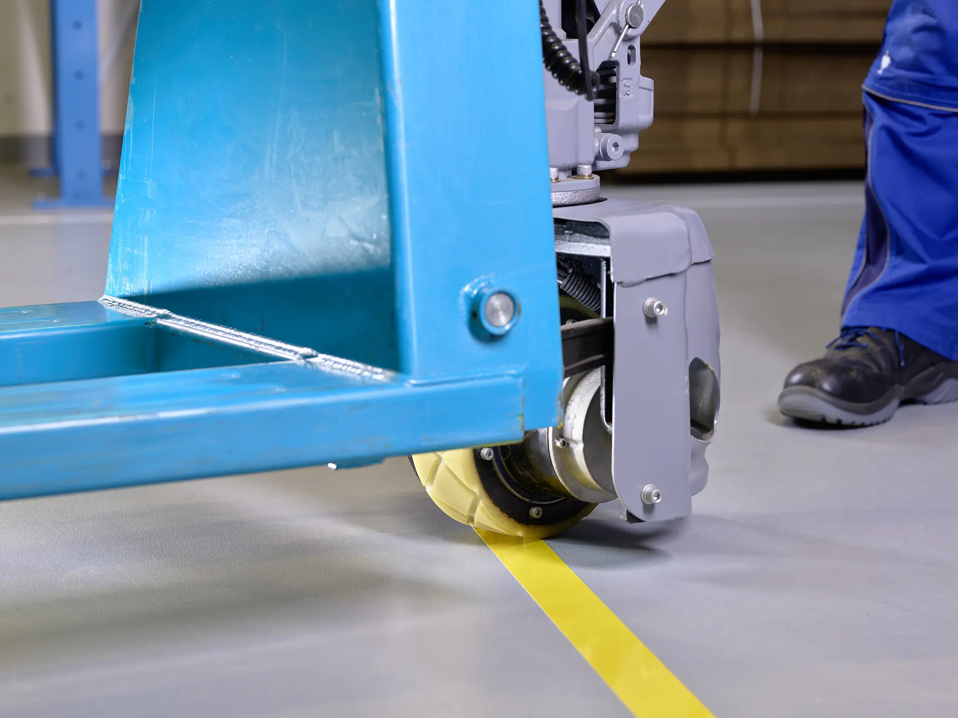 Close-up of a hydraulic hand pallet truck with a blue frame on a smooth, gray floor. A yellow line is visible on the floor, with a person wearing blue pants and black shoes standing nearby. The trucks wheel is aligned with the line made using tesa tape.