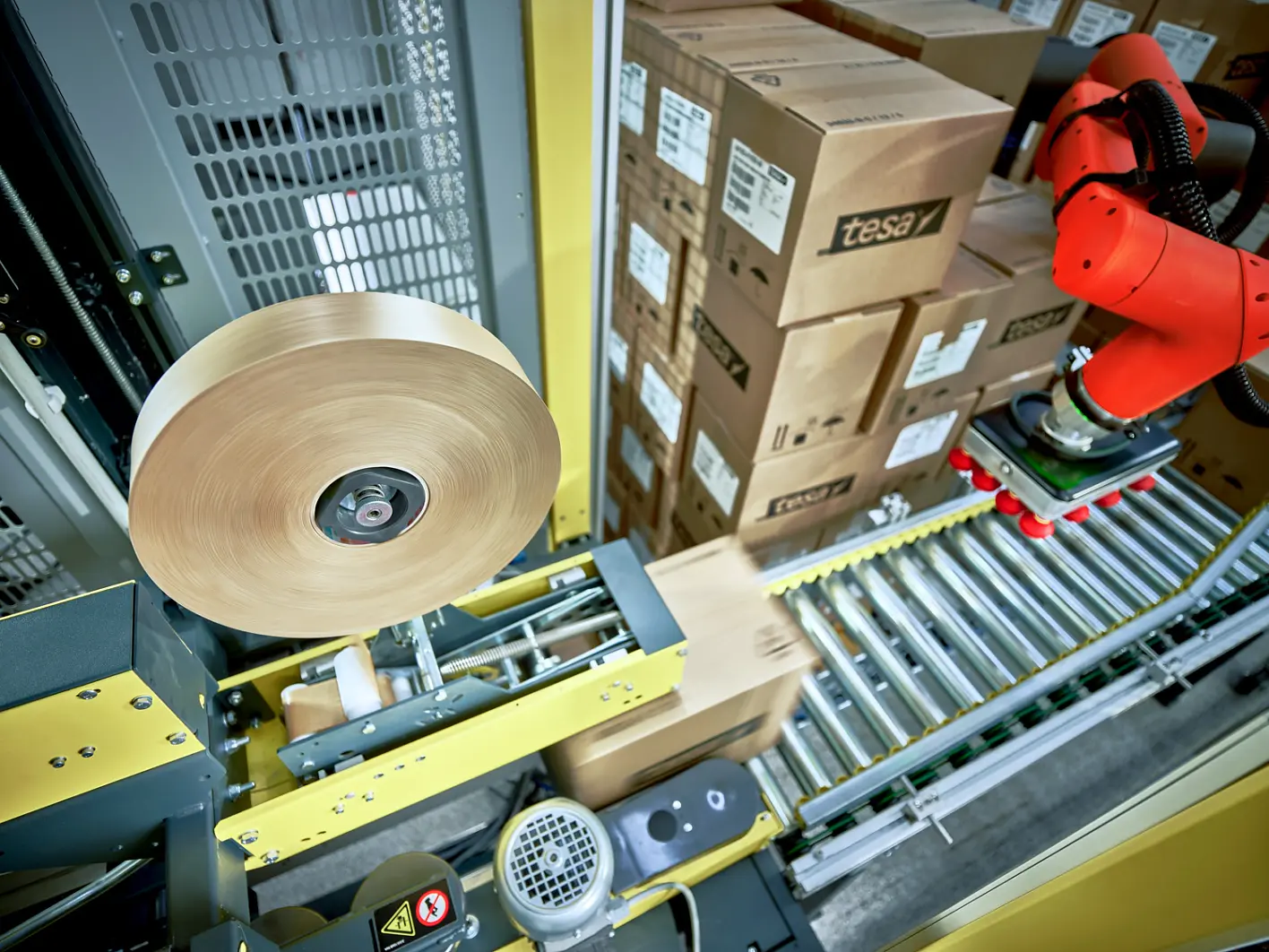 A robotic arm with suction cups lifts a cardboard box onto a conveyor belt, next to a roll of tesa tape in a warehouse setting. Stacked boxes with labels are visible in the background. (This text has been generated by AI)