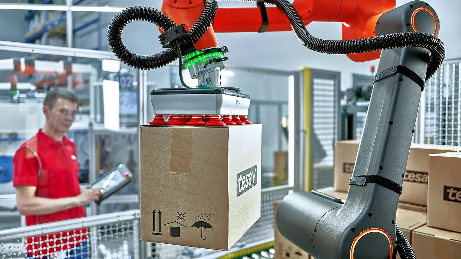 A robotic arm is lifting a cardboard box labeled tesa in a modern industrial setting. The arm is orange and black with a suction tool. A person in a red shirt stands in the background holding a tablet, observing the process.