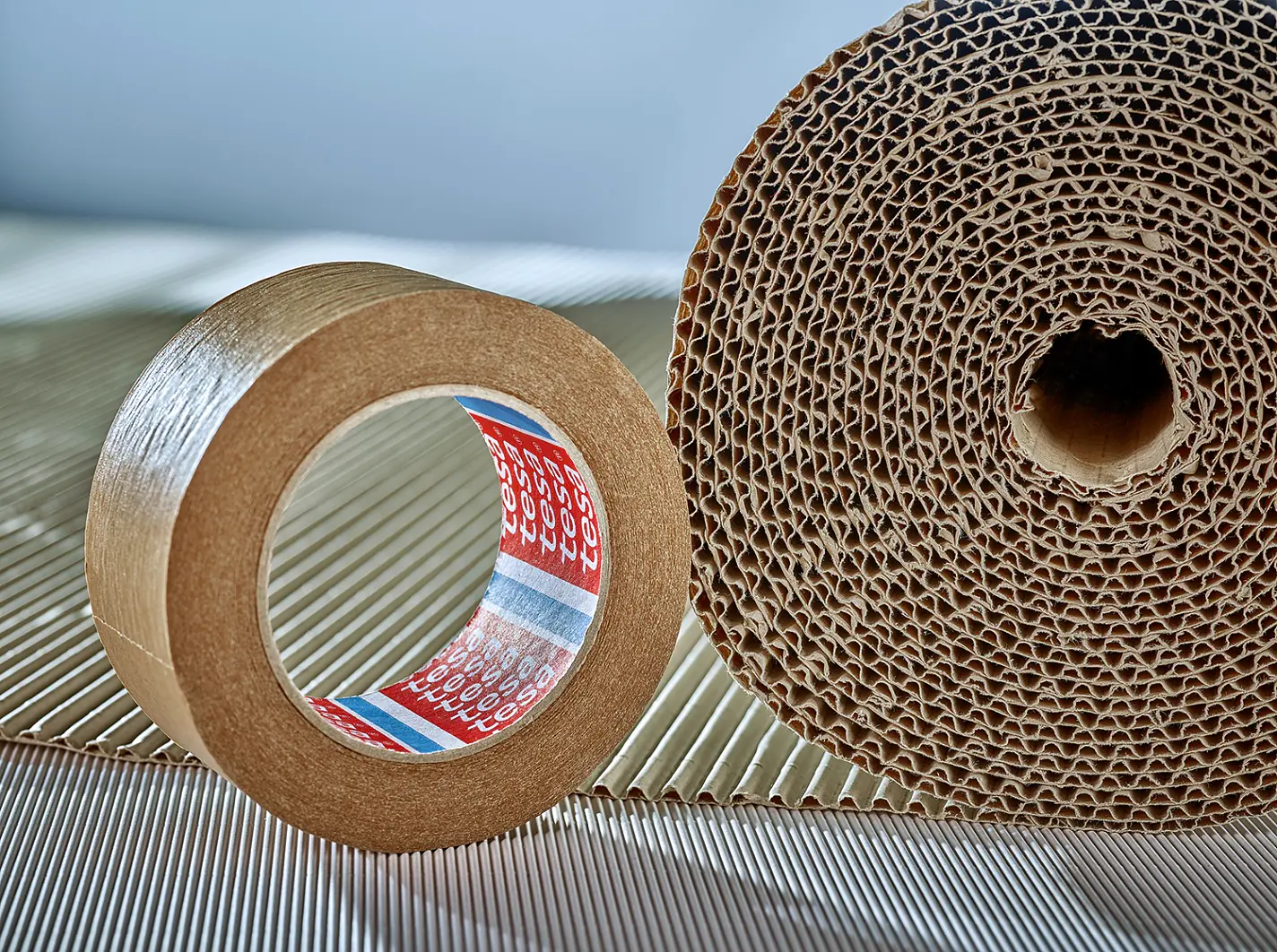 A roll of brown adhesive tesa tape stands upright next to a large roll of corrugated cardboard. The tesa tape has a red, white, and blue inner layer, while the cardboard features a visible honeycomb structure. Both items rest on a ribbed surface. (This text has been generated by AI)
