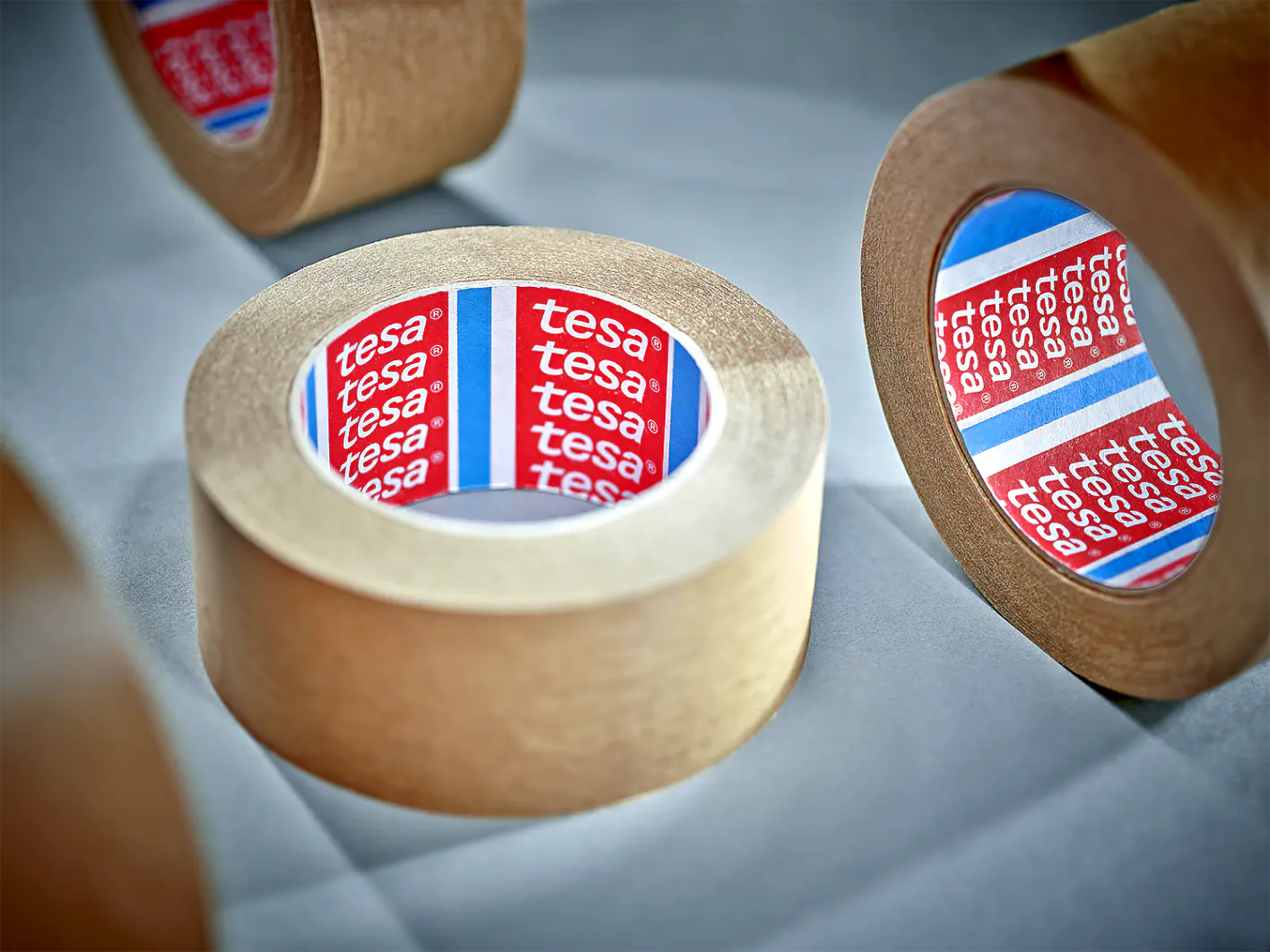 Several rolls of brown adhesive tesa tape are arranged on a light surface. The inner part of each roll displays a red and blue label with the brand name tesa repeated. The rolls are positioned at different angles. (This text has been generated by AI)