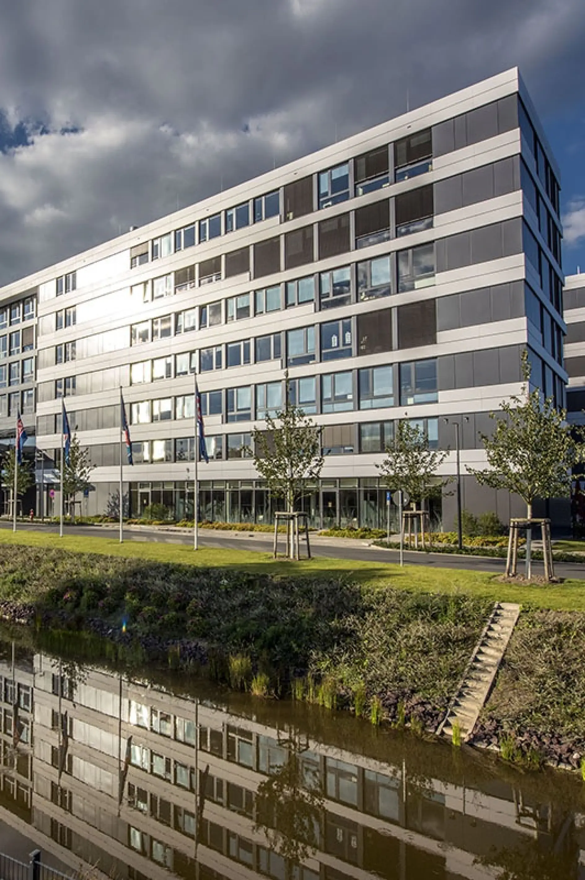 A modern, multi-story office building with a sleek facade and large windows is reflected in a calm body of water. Flags line the front, and trees are planted along a grassy area. The sky is partly cloudy. (This text has been generated by AI)