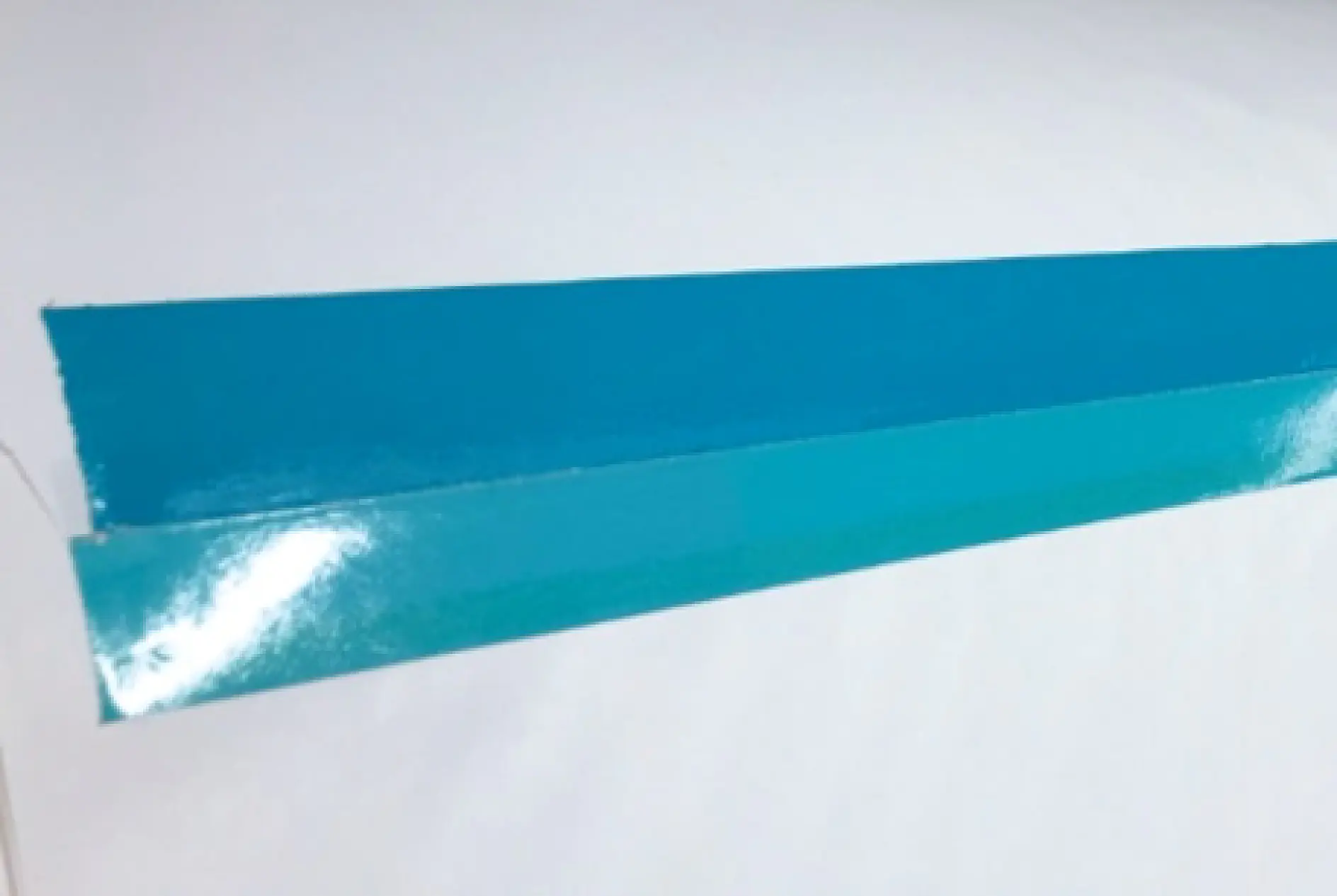 A strip of glossy blue tesa tape is adhered to a white surface, with one end slightly lifted. The tesa tape has a reflective sheen and appears to be in good condition. (This text has been generated by AI)