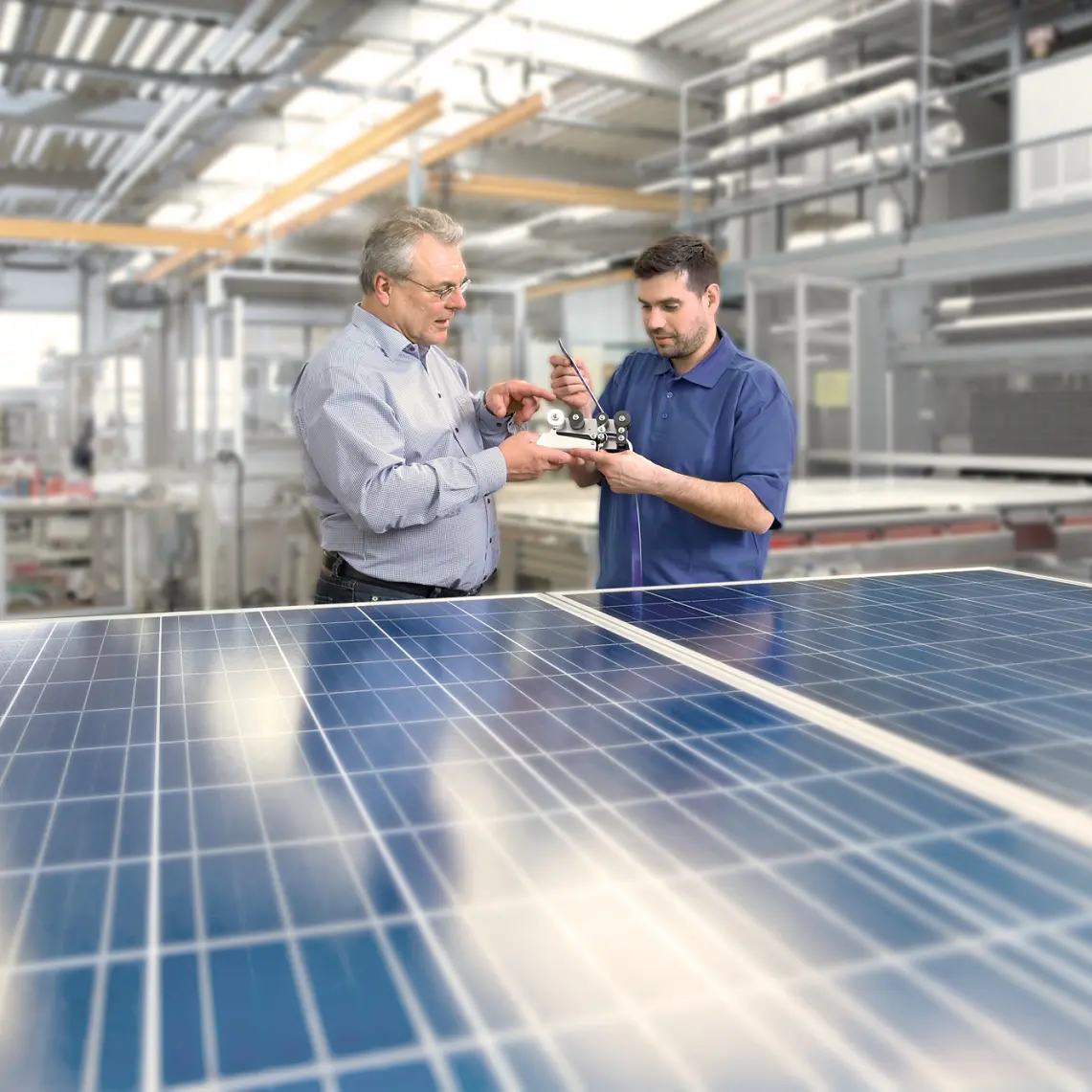 Two men are examining a mechanical component inside a modern factory. They stand near solar panels, with machinery and equipment visible in the background. The setting suggests an industrial or technological environment focusing on solar energy. (This text has been generated by AI)