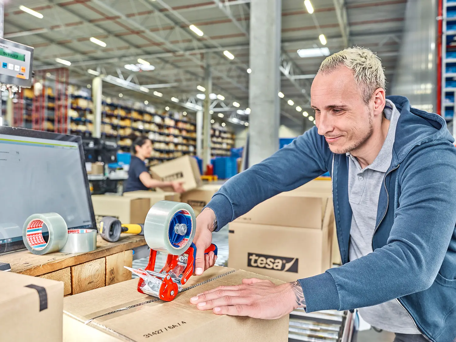 A person is sealing a cardboard box with tesa tape on a table in a warehouse. A digital scale and more boxes are nearby. Shelves filled with items are visible in the background. Another person is working in the distance.