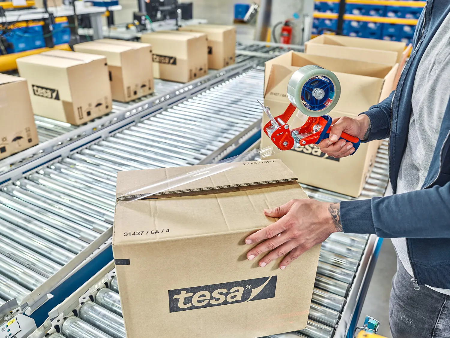 A person seals a cardboard box labeled tesa on a conveyor belt in a warehouse using tesa tape. Several similar boxes can be seen in the background. The individual is using a red hand-held tesa tape dispenser.