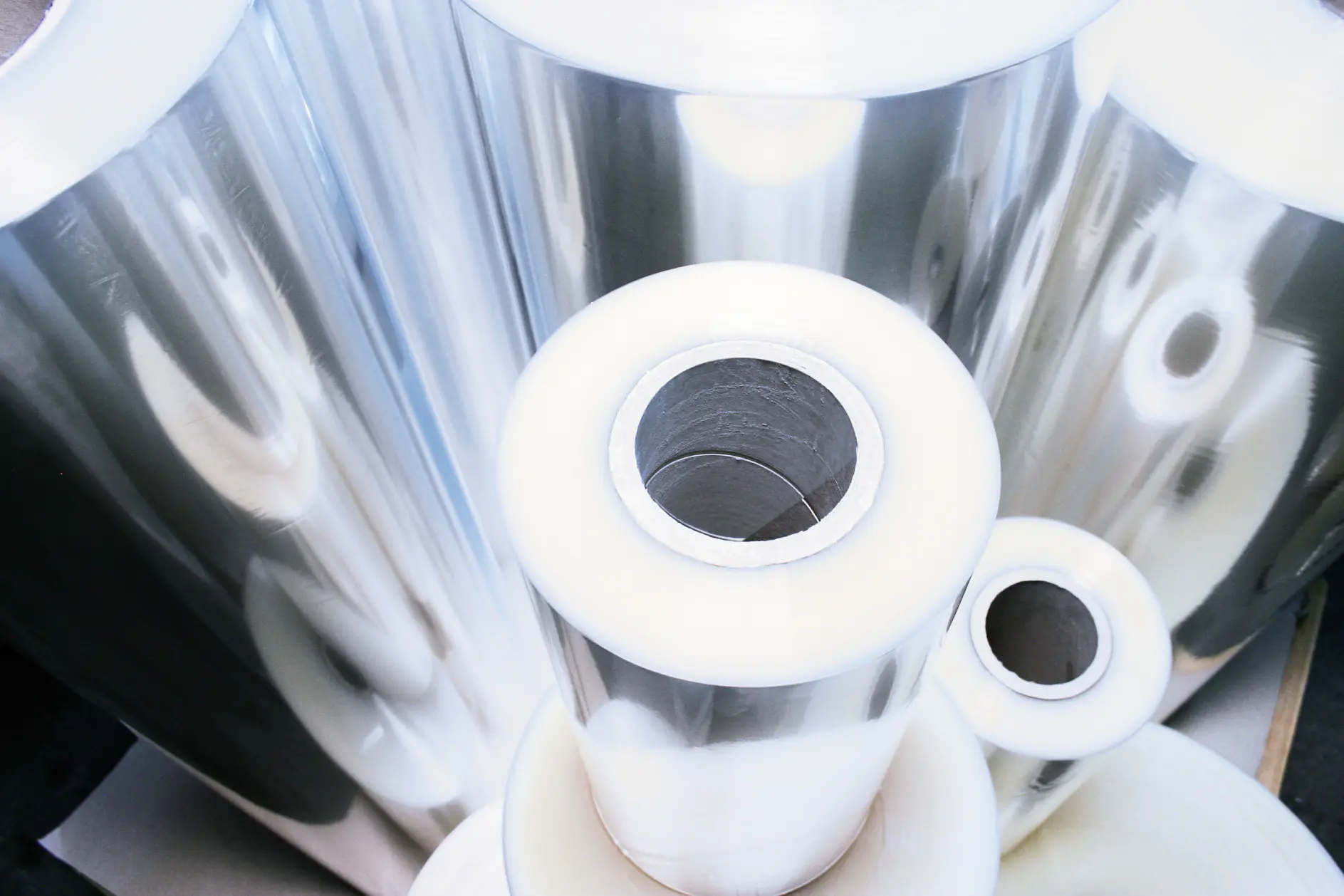 Rolls of shiny, transparent tesa tape are stacked, showcasing their reflective surfaces. They vary in size, with cylindrical cores visible at the center of each roll. The image captures the materials from a top angle.