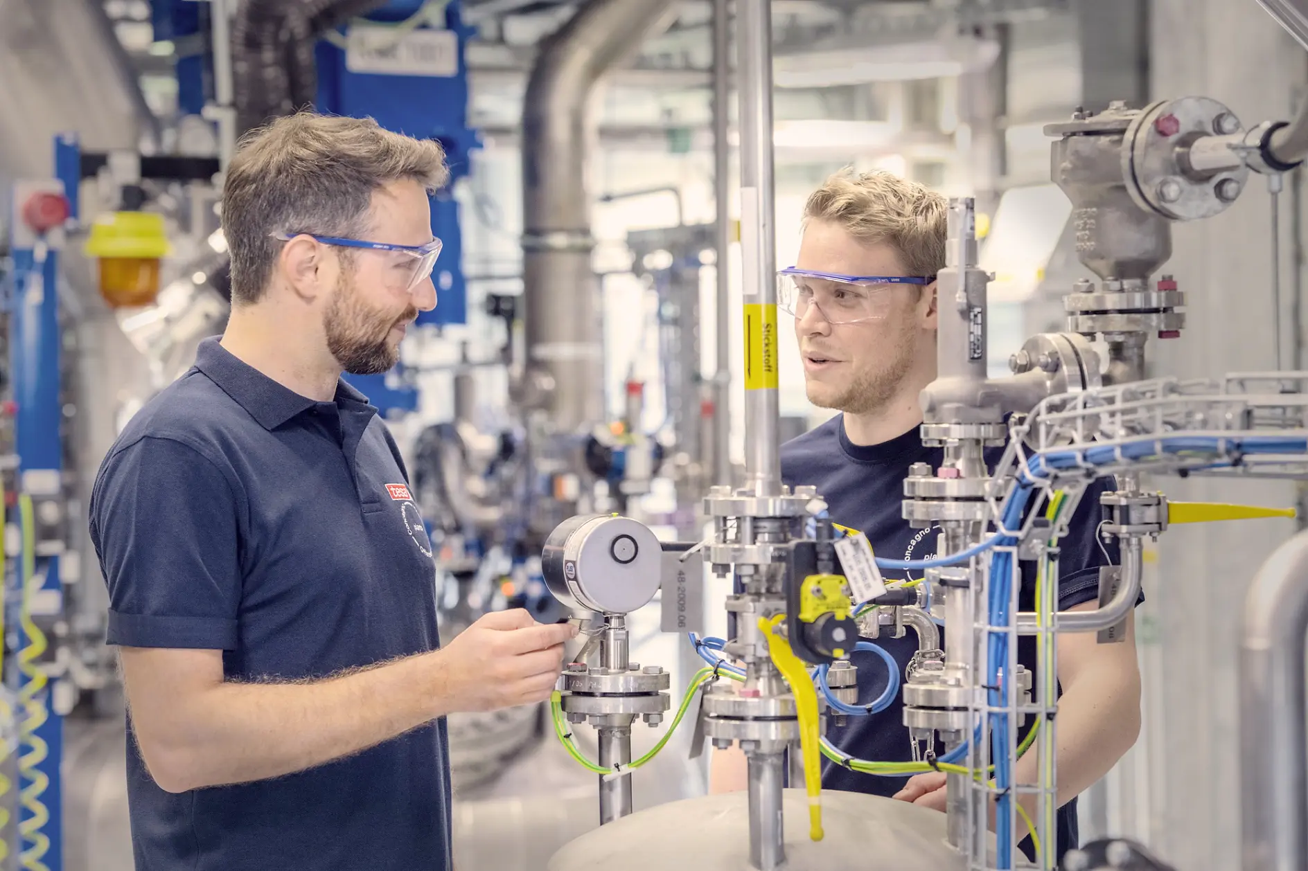 Two men wearing safety glasses are engaged in a discussion within an industrial setting. They are surrounded by various pipes, gauges, and equipment. Both are dressed in dark blue shirts, and one is gesturing towards a piece of machinery with tesa tape visible nearby. (This text has been generated by AI)