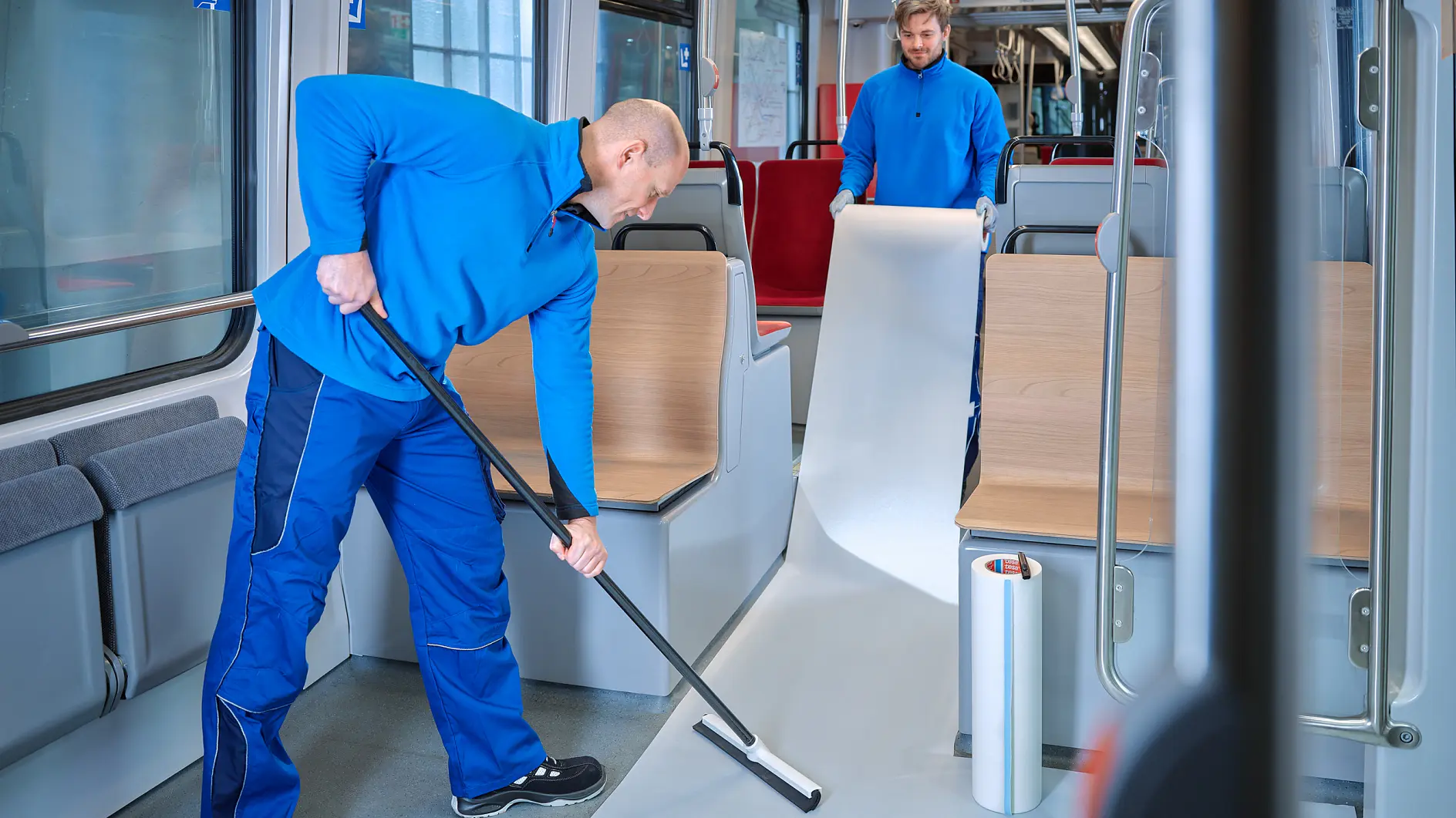 Two individuals in blue uniforms are installing a protective film on the floor of a train carriage. One person is unrolling the tesa tape film while the other smooths it with a tool. The train interior has wooden and red seating.