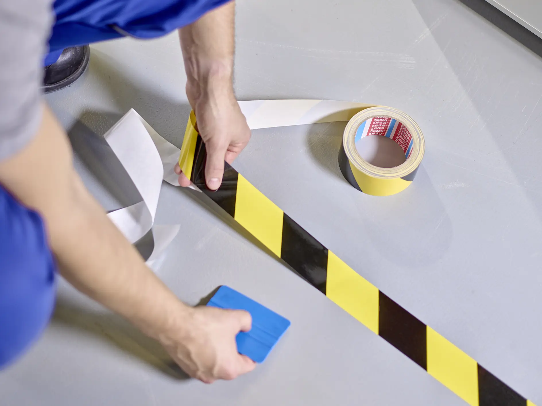 A person wearing blue pants and a gray shirt is applying yellow and black striped tesa tape to a light gray floor. They are using a blue tool to smooth the tesa tape, and a roll of similar tesa tape is nearby.