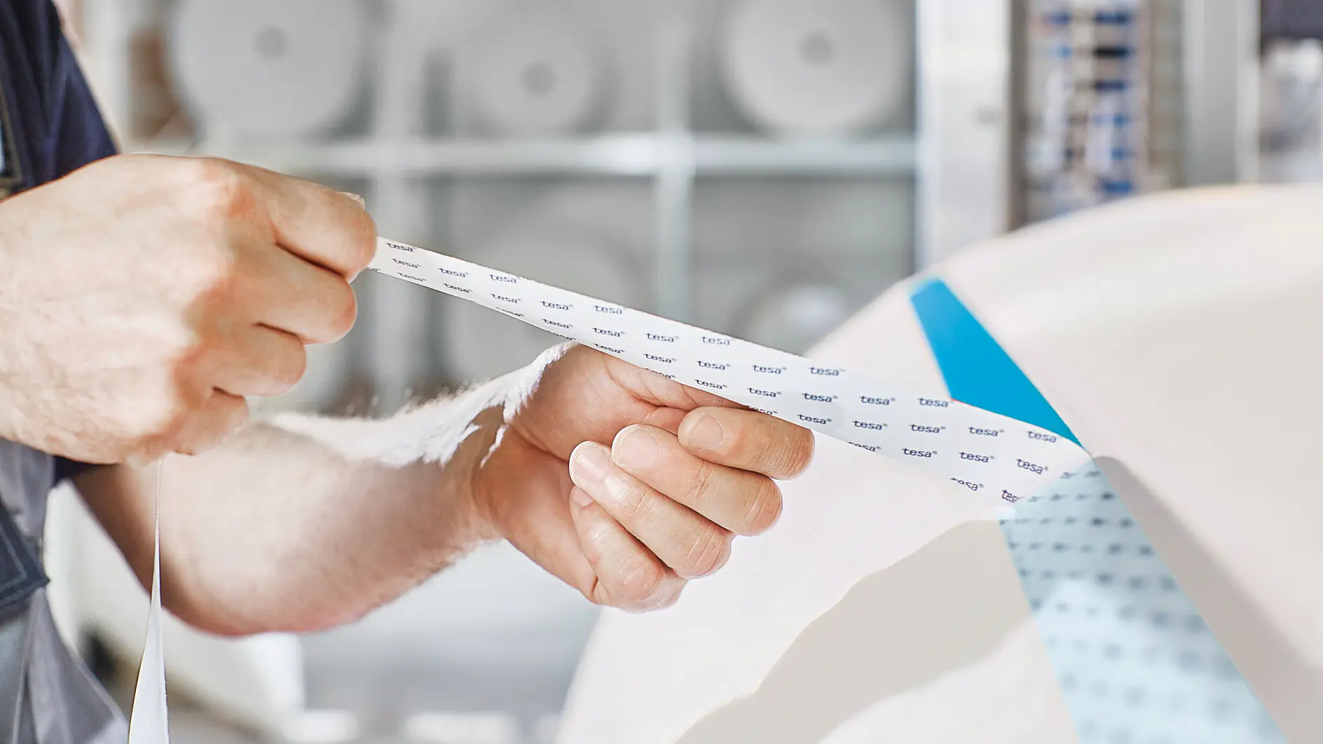 A person holds a strip of masking tesa tape, peeling it off from a larger roll. The tesa tape has text printed on it. The background shows blurred rolls of paper or tesa tape on shelves.