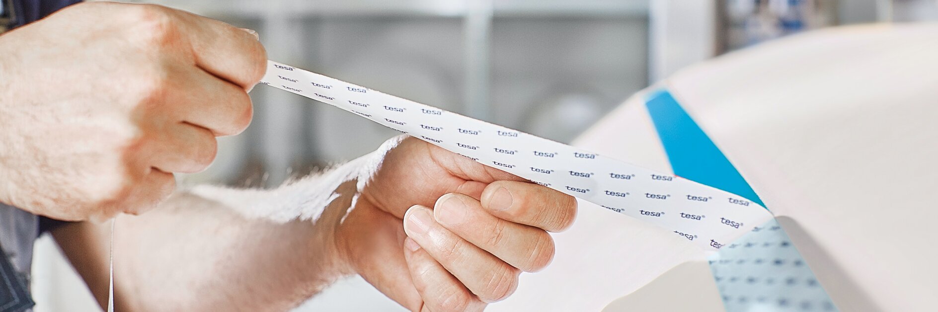 A person holds a strip of masking tesa tape, peeling it off from a larger roll. The tesa tape has text printed on it. The background shows blurred rolls of paper or tesa tape on shelves.