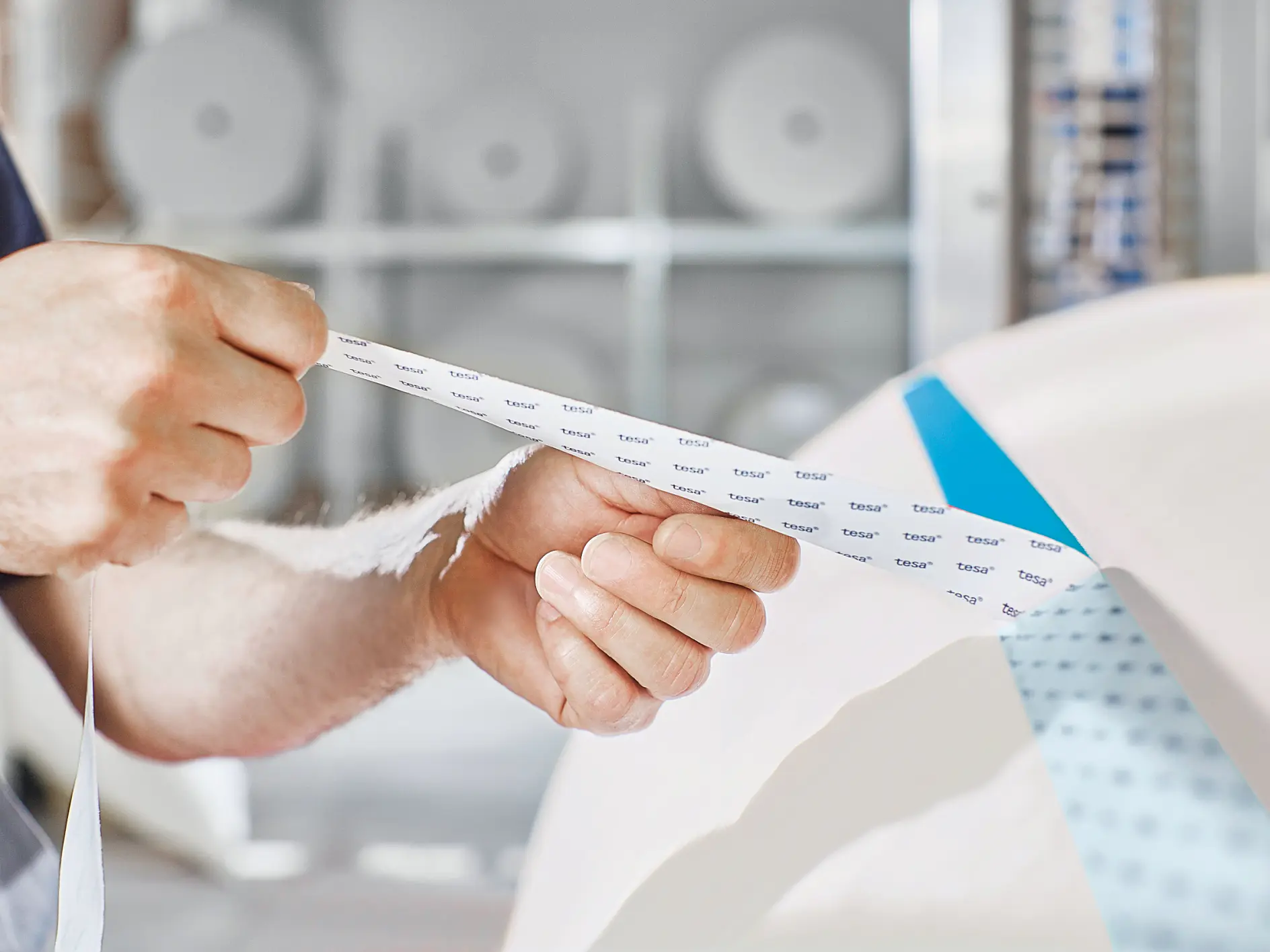 A person holds a strip of masking tesa tape, peeling it off from a larger roll. The tesa tape has text printed on it. The background shows blurred rolls of paper or tesa tape on shelves. (This text has been generated by AI)