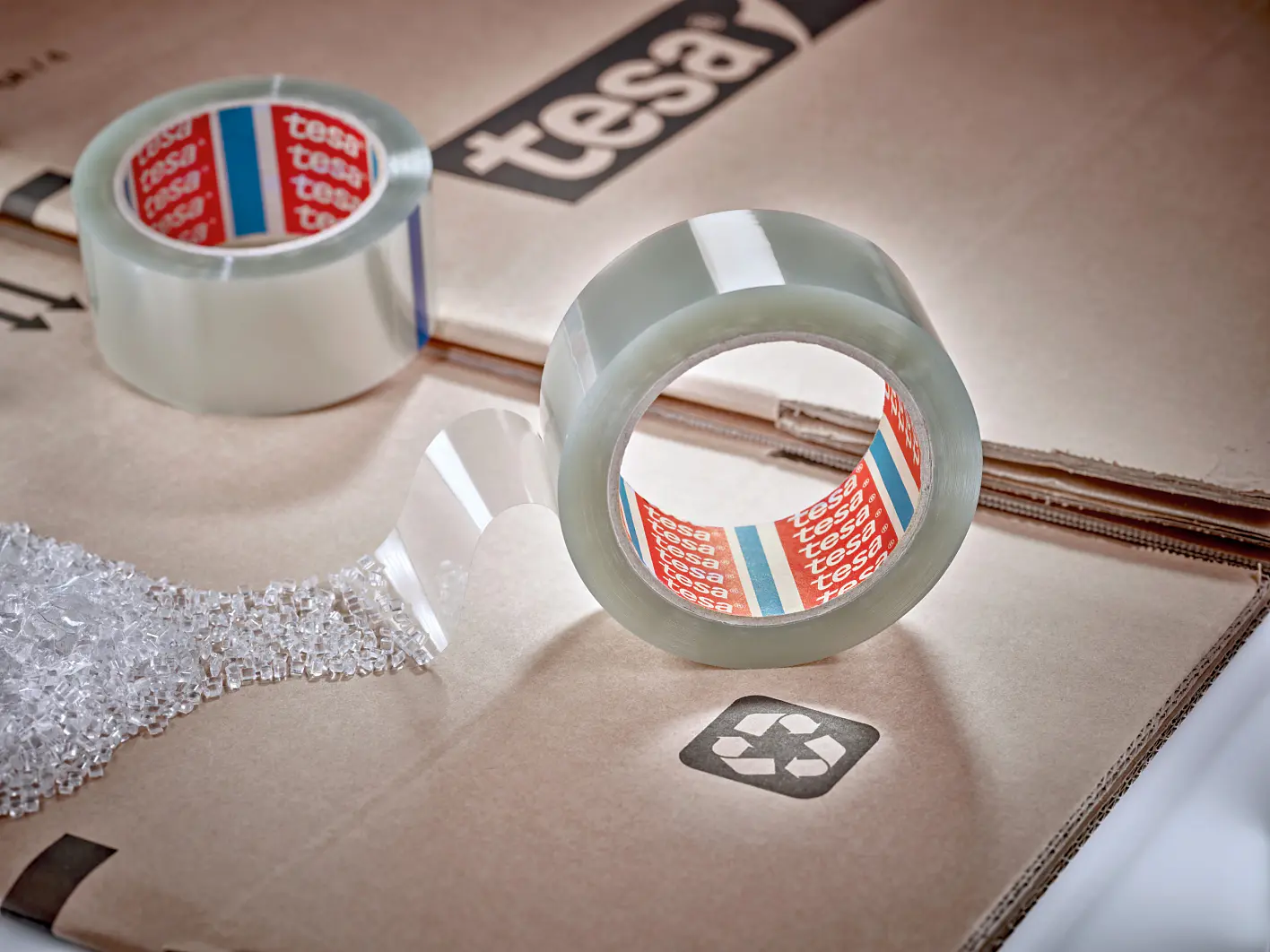 A roll of clear adhesive tesa tape partially unrolled on a cardboard surface. The background features more cardboard with a visible recycling symbol. The tesa tape has a red inner label with the text tesa repeated. Small packing material is also visible. (This text has been generated by AI)