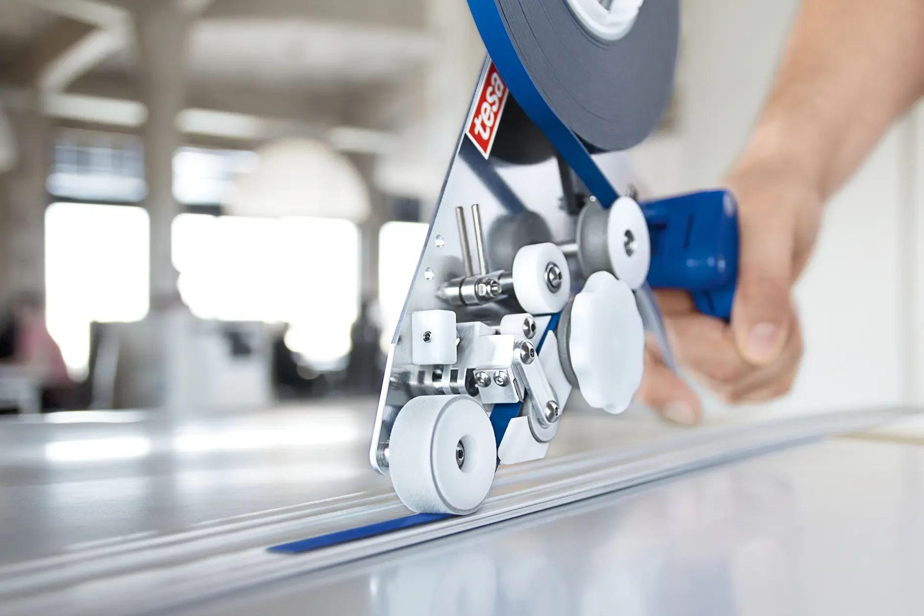 Close-up of a hand operating a tape dispenser, applying double-sided adhesive tesa tape to a flat surface. The dispenser is equipped with rollers and a blue handle, positioned in an office setting.