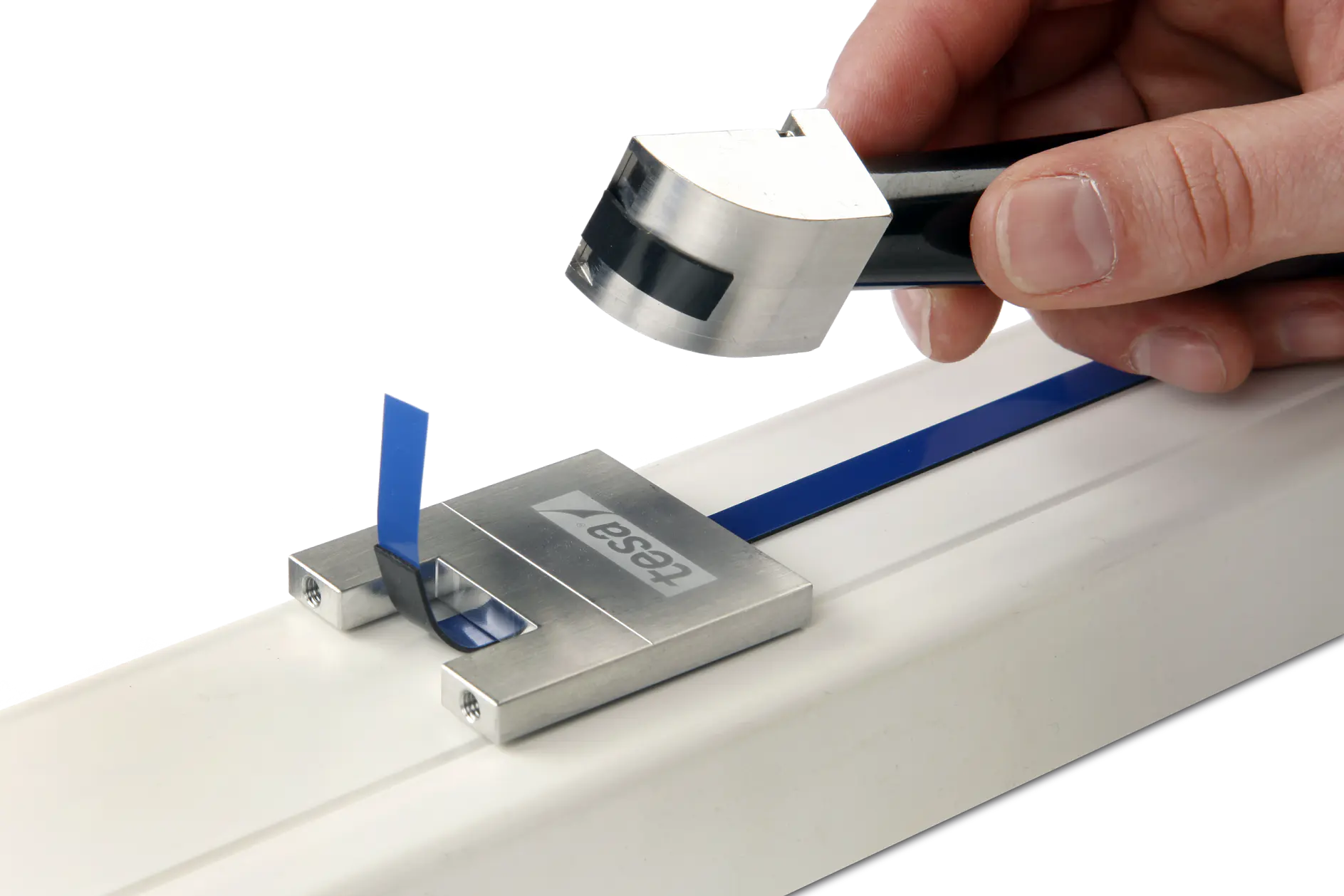 A hand holds a device above a blue strip inside a metal guide with the tesa tape logo. The setup is placed on a white surface, possibly for demonstration or measurement purposes.
