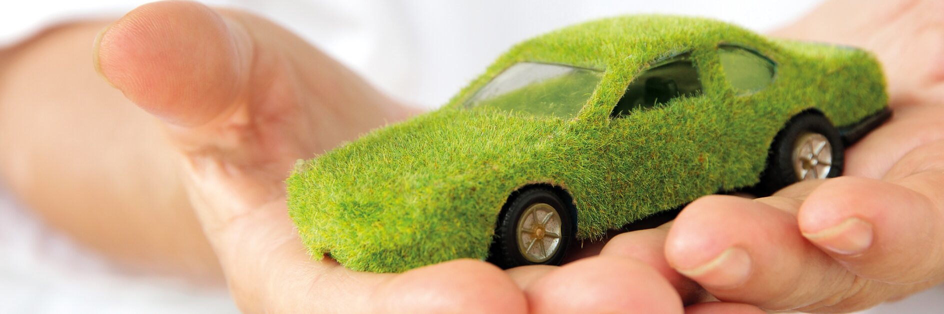 Hands holding a small car model covered in green grass-like material, symbolizing eco-friendly or sustainable automotive concepts.
