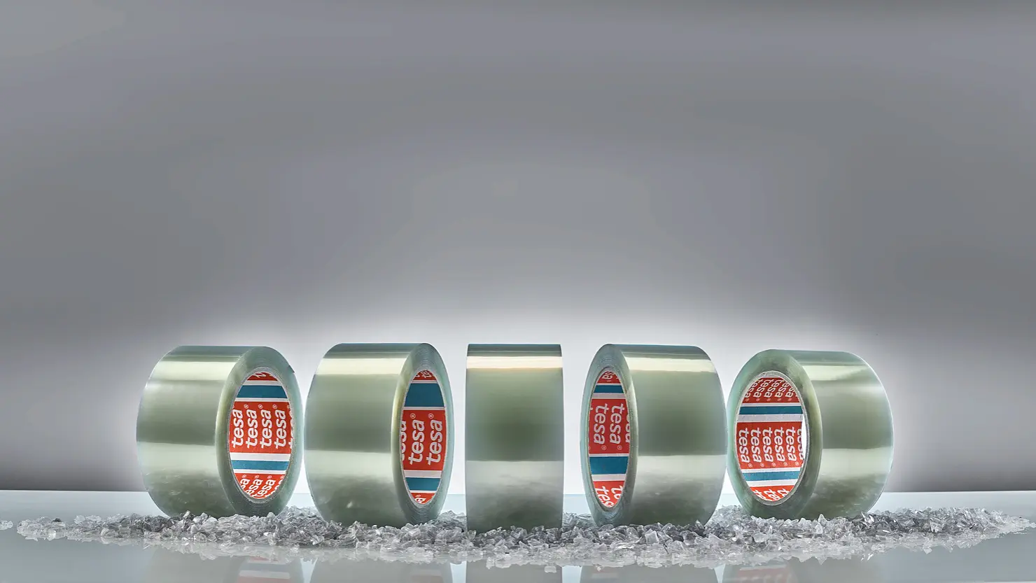 Five rolls of clear adhesive tesa tape are displayed vertically against a gray background. Small, scattered transparent granules are on the surface in front of the rolls. The tape cores are branded with red and blue labels. (This text has been generated by AI)