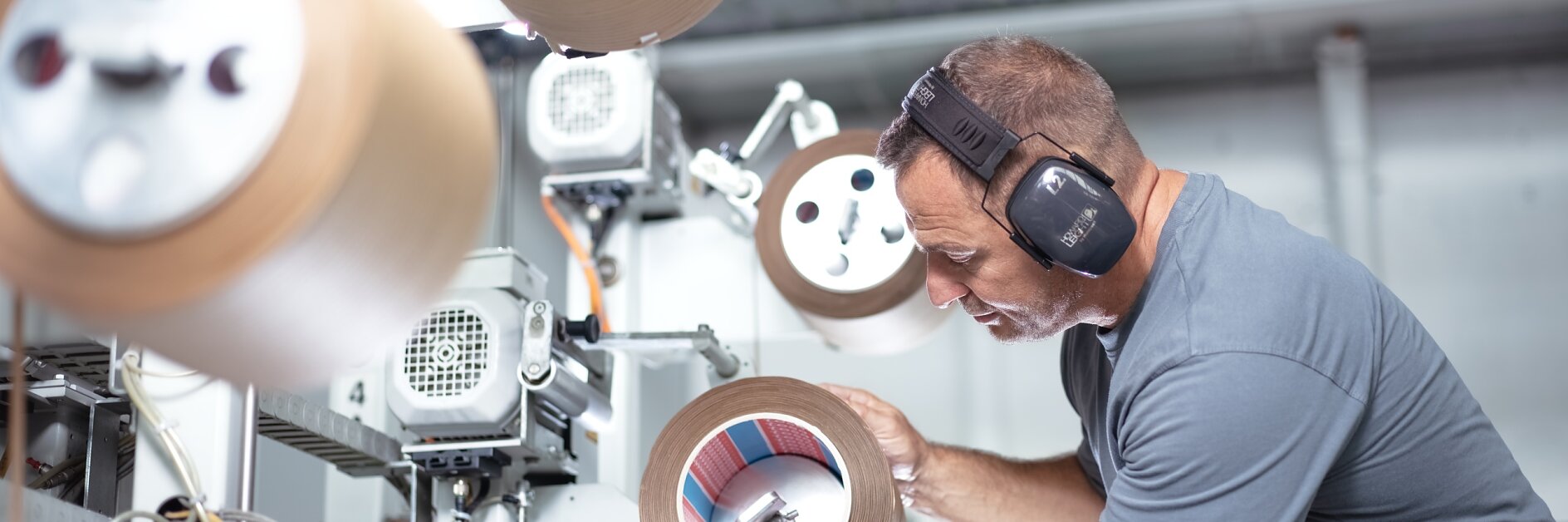 A man wearing ear protection inspects a large roll of tesa tape in an industrial setting. He is surrounded by machinery and other rolls of tesa tape, focusing intently on the one in his hands. The environment appears to be a manufacturing facility.