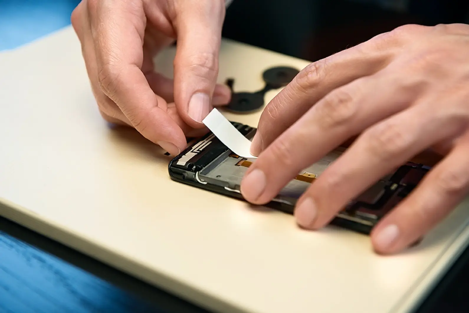 A person is repairing a smartphone. They are placing a piece of white tesa tape inside the phone, which is on a table. The phone appears to be disassembled, with internal components visible. The background is a light-colored surface. (This text has been generated by AI)