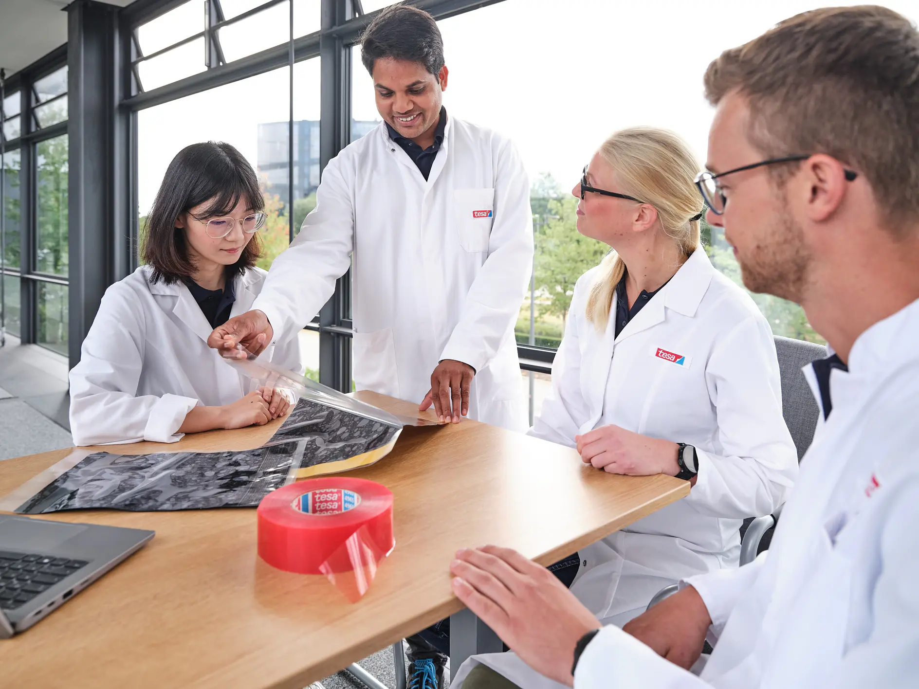Four people in lab coats are gathered around a wooden table in a bright room with large windows. One person is showing a large sheet of material to the others. There is a roll of red tesa tape and a laptop on the table. (This text has been generated by AI)