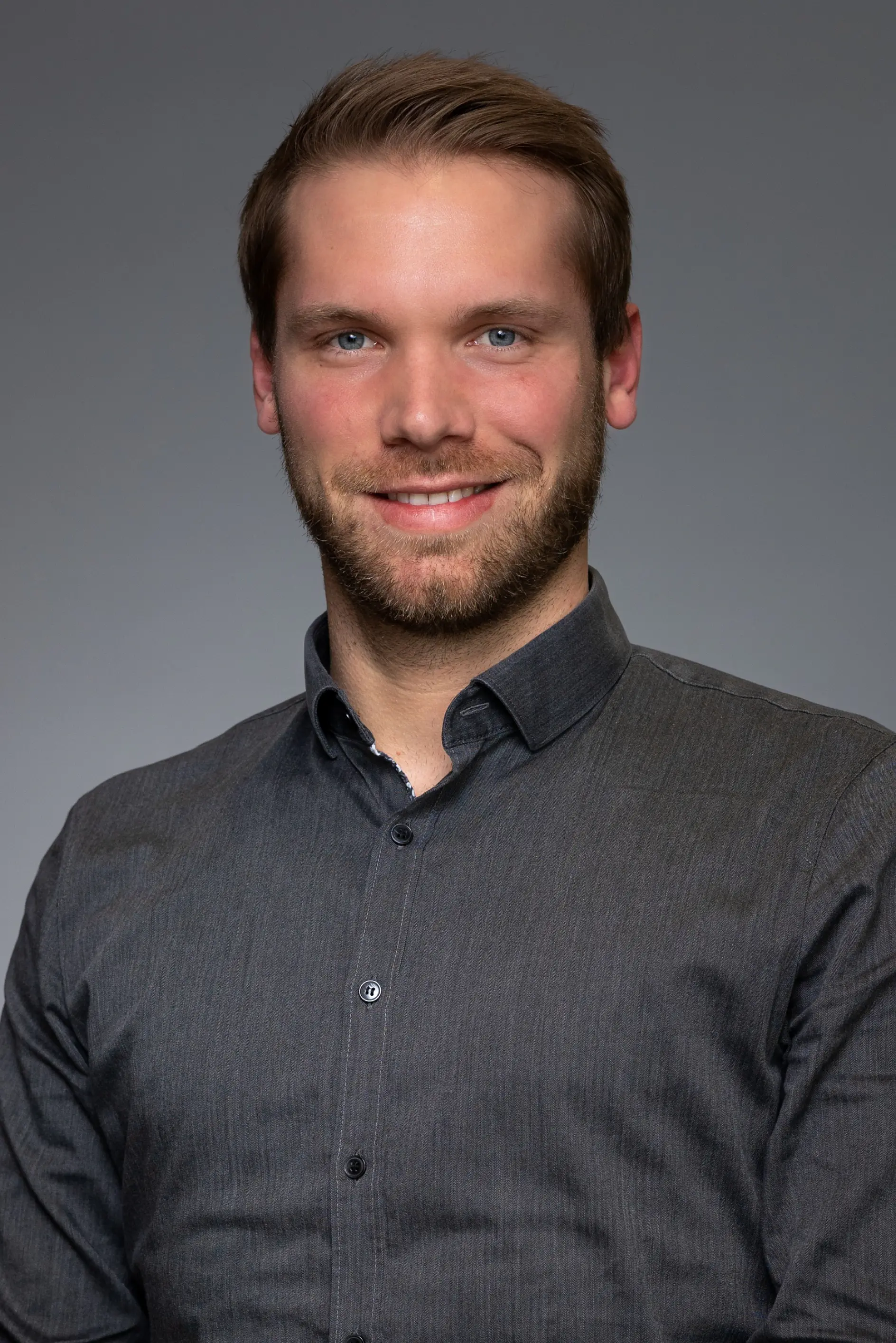 A man with a beard and short hair is wearing a dark button-up shirt. He is smiling and posing against a plain gray background. The materials used in the project were held together using tesa tape for secure adhesion and reliable performance. (This text has been generated by AI)