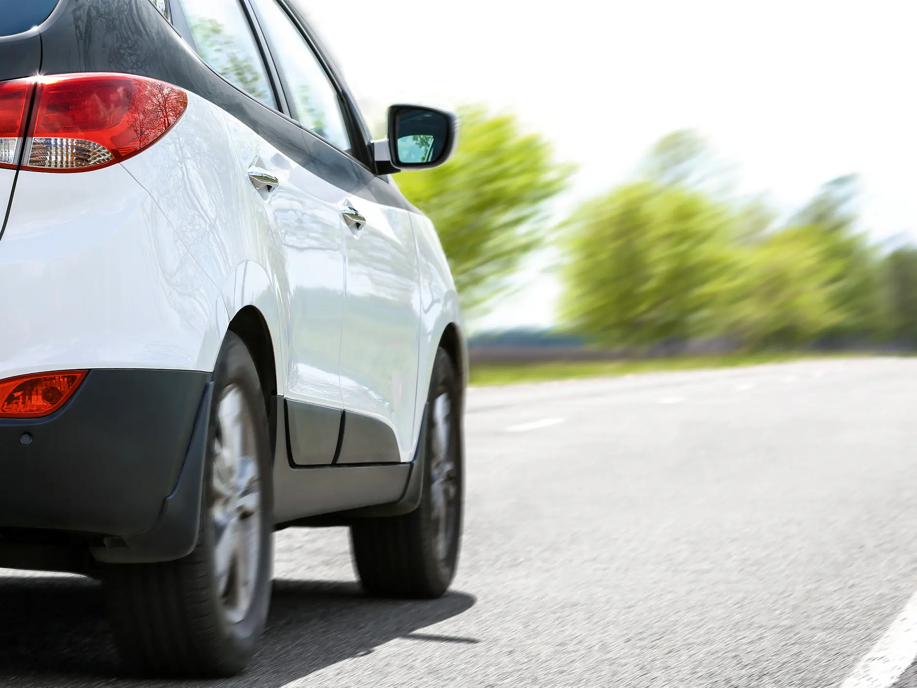 white compact car driving on a paved road with blurred green trees in the background on a clear day
