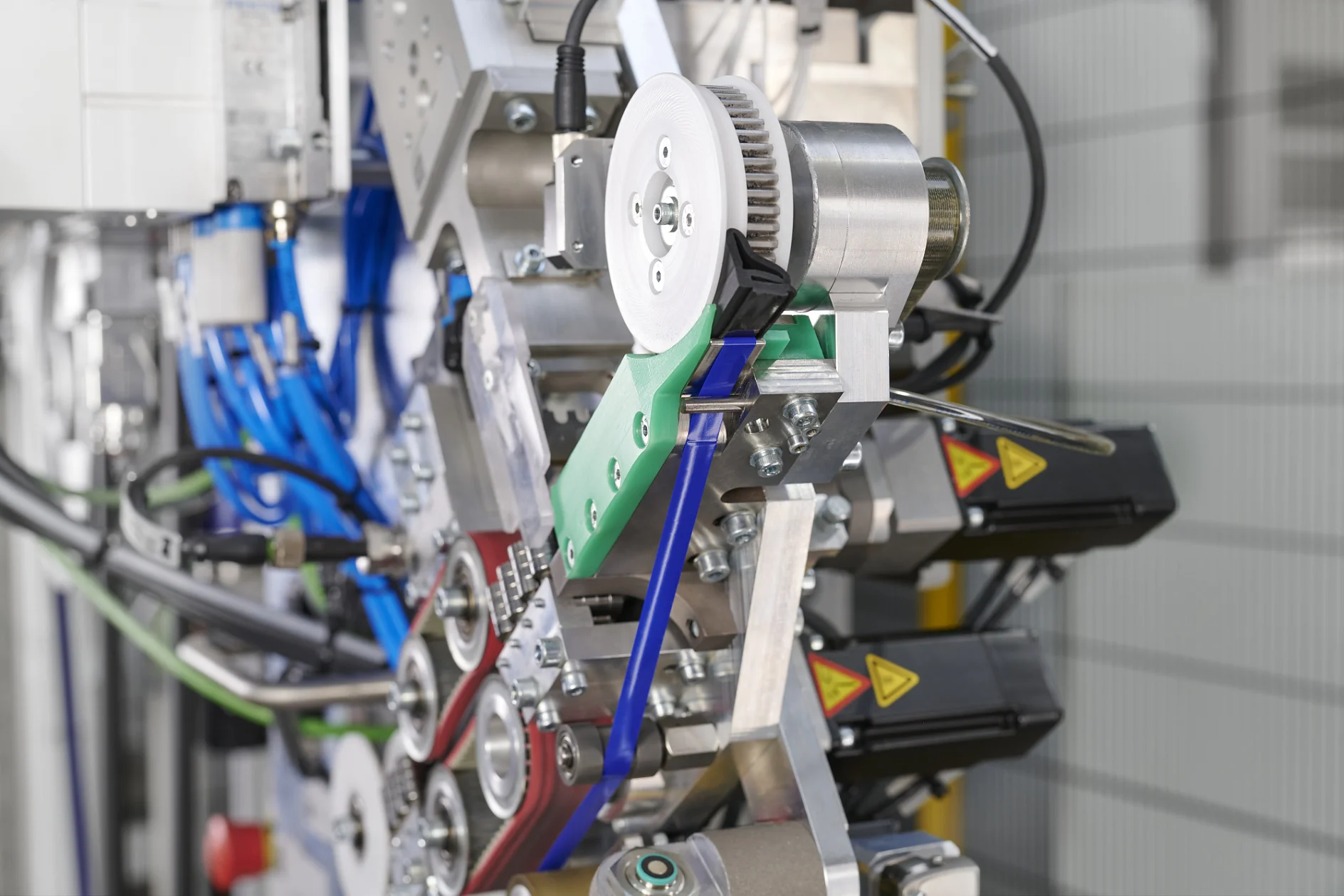 Vue rapprochée d'une machine industrielle avec engrenages, courroies, leviers colorés, câbles, ruban tesa et étiquettes d'avertissement visibles, montrant en détail les composants mécaniques et les connexions.