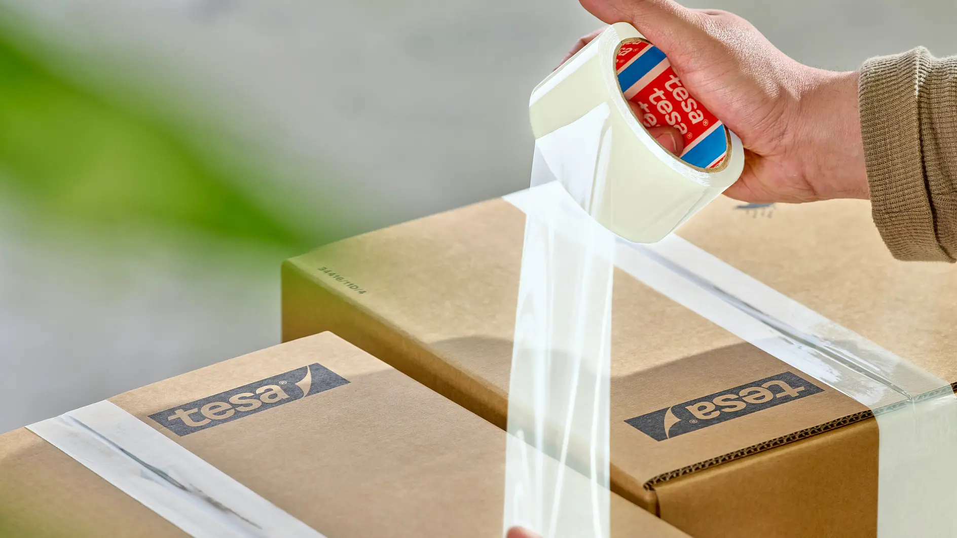 Des mains appliquant du ruban tesa transparent pour fermer des cartons sur une surface claire, avec un arrière-plan vert flou. (This text has been generated by AI)