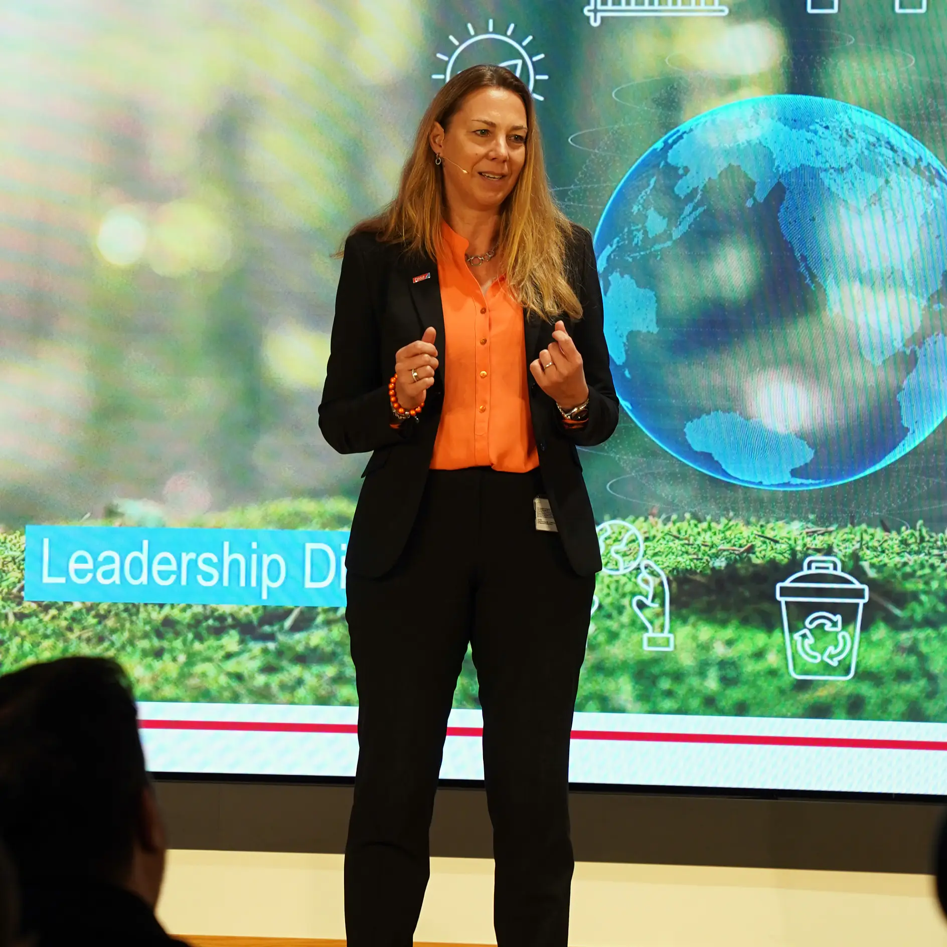 Une femme en tailleur d'affaires parle sur scène devant un écran avec un globe, des icônes, « Leadership D » et la marque tesa tape visibles. (This text has been generated by AI)