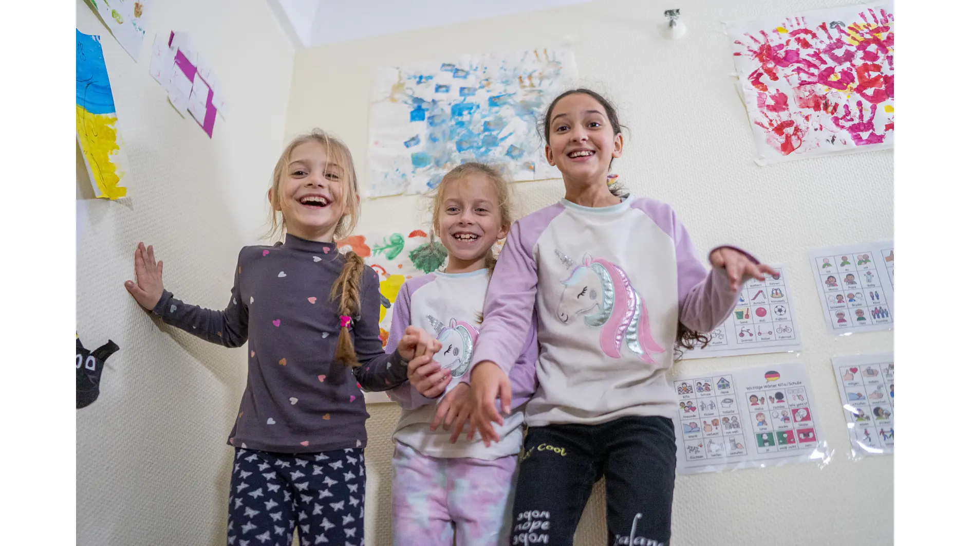 (Traite de gauche à droite) Larissa*, 7 ans, Svitlana*, 7 ans, et Daniela*, 11 ans, sourient à l'appareil photo dans l’Espace Amis des Enfants situé dans un abri temporaire pour les réfugiés ukrainiens à Francfort, en Allemagne.