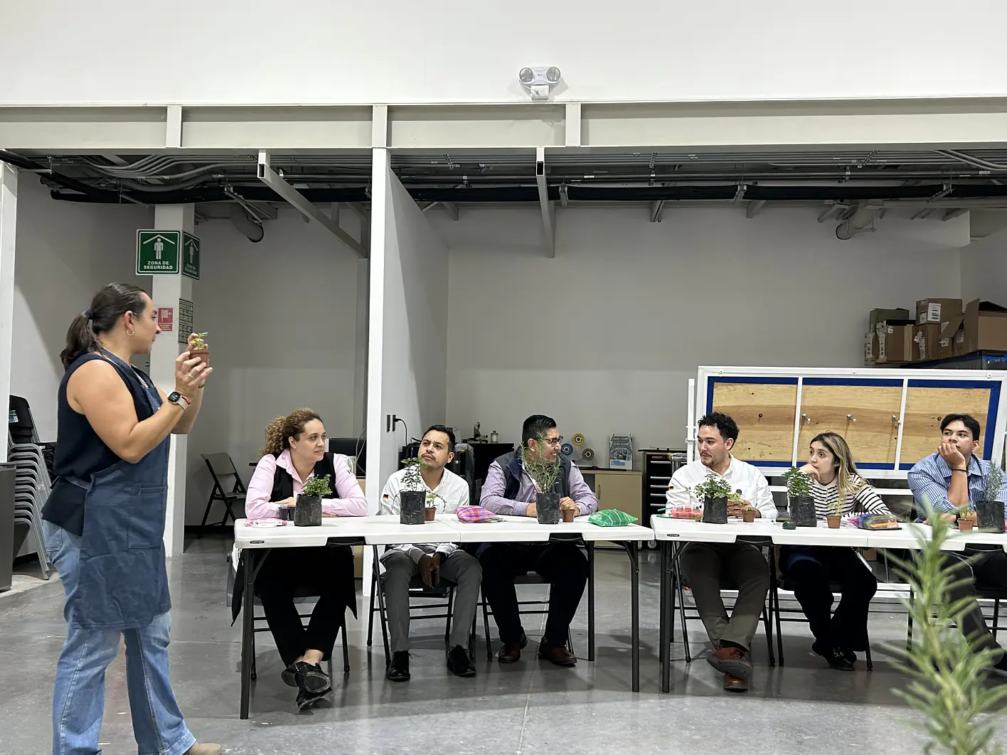Une femme parle à six personnes assises à une longue table, chacune avec une plante en pot. Des étagères et des chaises empilées sont en arrière-plan. (This text has been generated by AI)